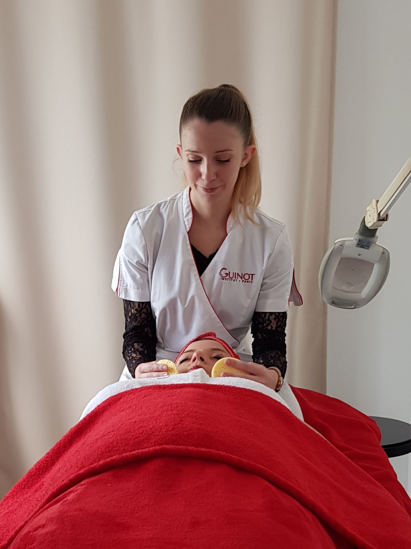 Une esthéticienne pratiquant des soins sur le visage d'une cliente allongée et recouverte d'une couverture rouge