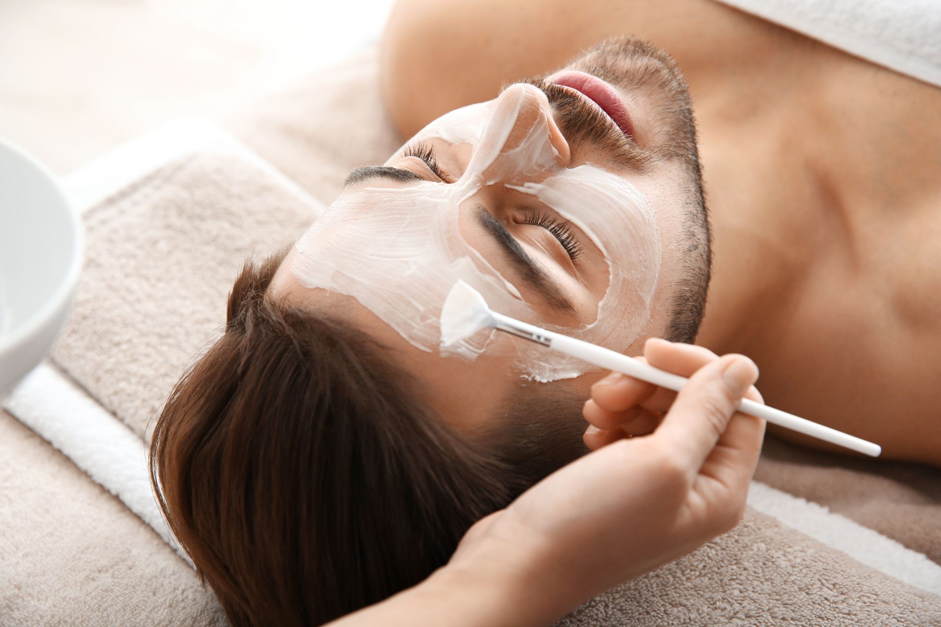 Application d’un masque crème sur le visage d’un homme allongé, à l'aide d'un pinceau
