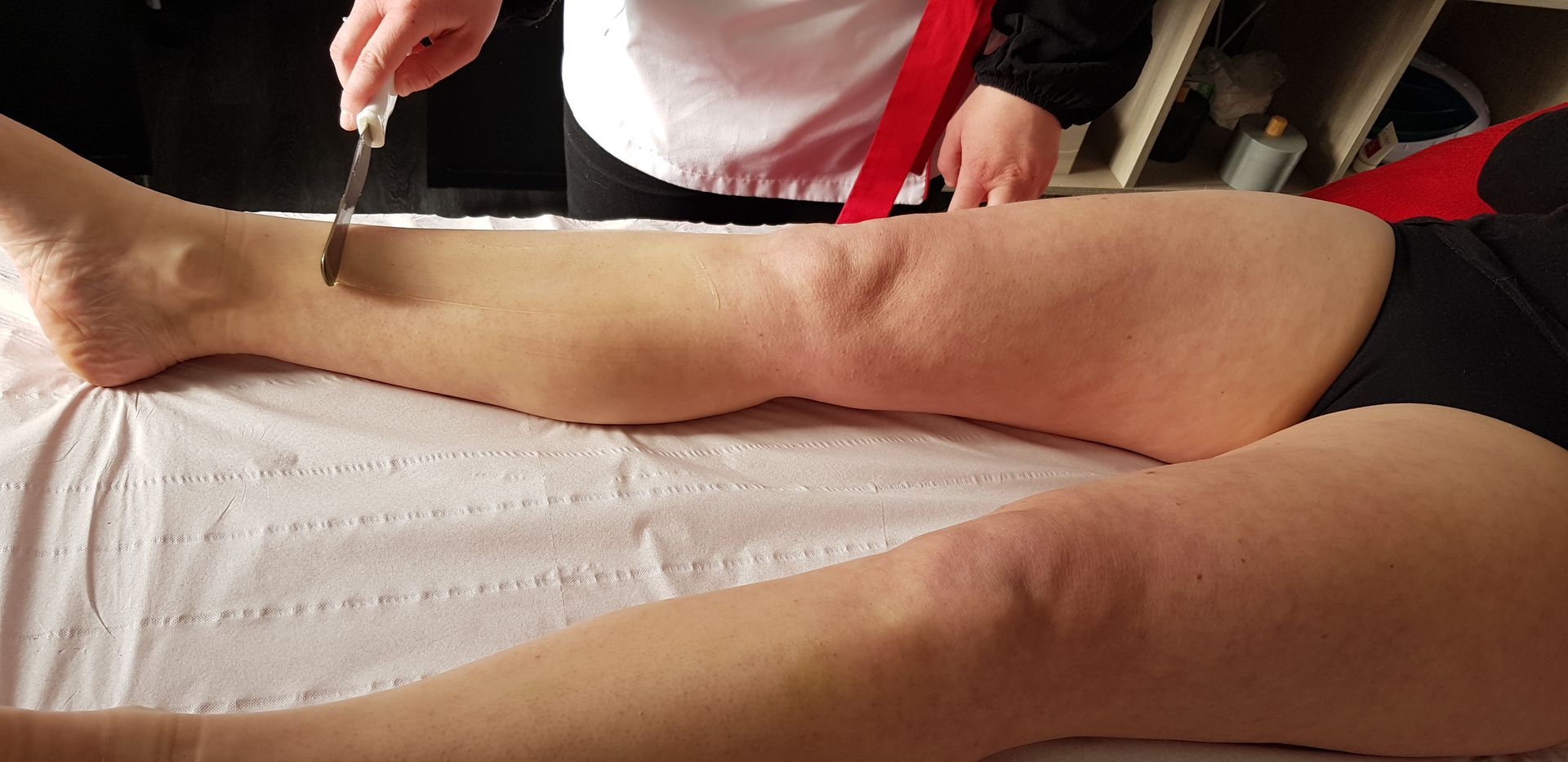 Application de cire tiède à la spatule sur la jambe d’une cliente allongée pour une épilation en institut