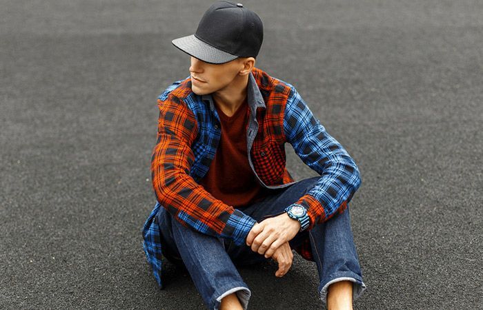 Mustaan lippalakkiin, flanellipaitaan ja farkkuihin pukeutunut henkilö istuu asfaltilla ja katsoo sivuun.