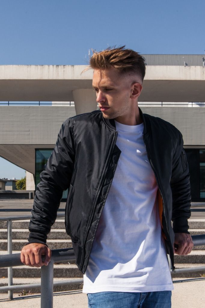 Mustaan ​​bombertakkiin ja valkoiseen paitaan pukeutunut mies katsoo poispäin ja nojaa metallikaiteeseen.