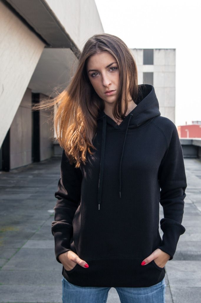 Mustaan ​​huppariin ja farkkuihin pukeutunut nainen seisoo ulkona kädet taskuissa ja katsoo kameraan.