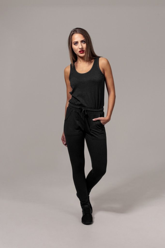 Nainen mustassa haalarissa, kädet taskuissa, katsoo kameraa harmaata taustaa vasten.