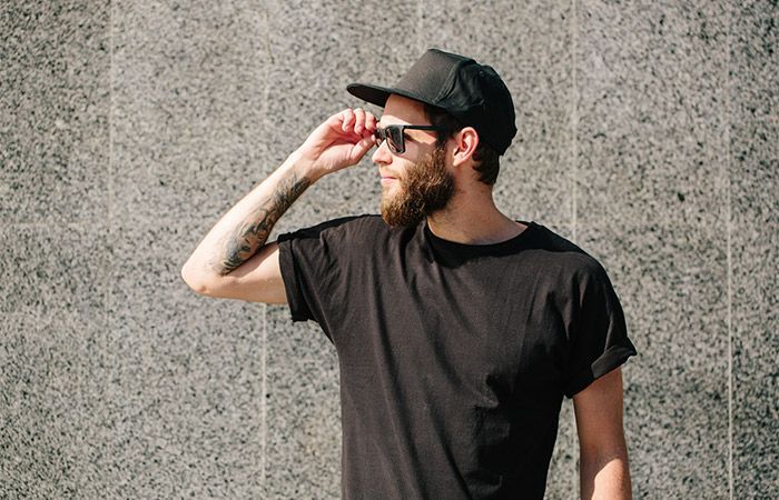 Mustassa lippalakissa ja aurinkolaseissa oleva mies katsoo oikealle, käsi silmälaseilla, harmaata seinää vasten.