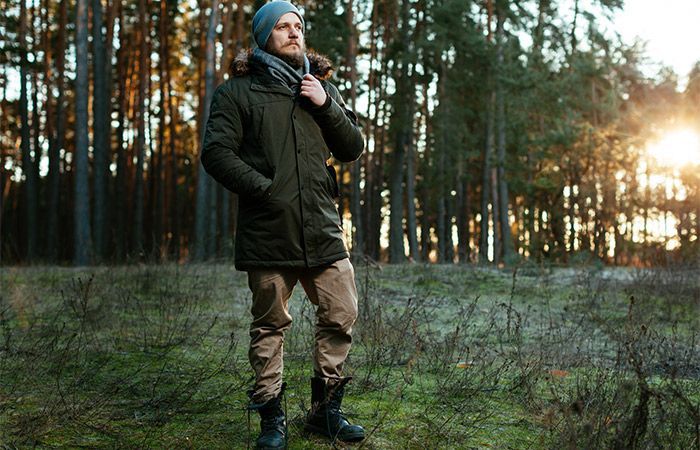 Vihreässä takissa ja hatussa oleva mies seisoo metsässä, auringonvalo paistaa puiden läpi.