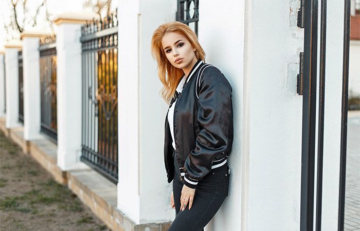 Nainen nojaa valkoista seinää vasten, yllään musta takki, mustat housut ja valkoinen paita, lähellä aitaa.