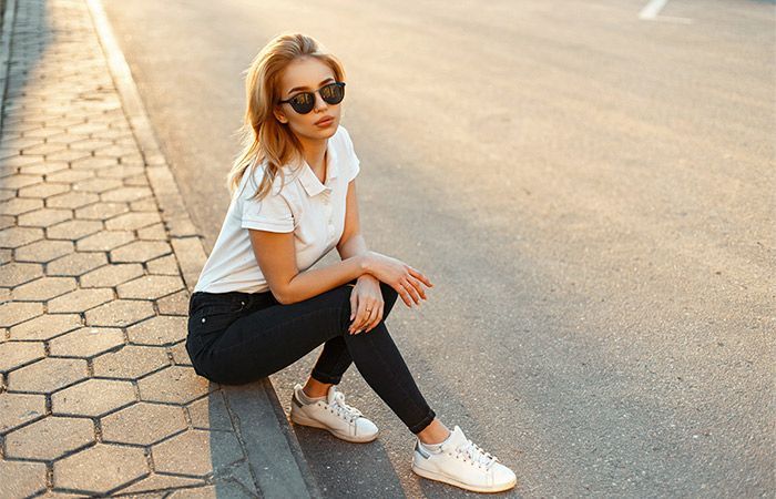 Aurinkolaseissa oleva nainen istuu jalkakäytävällä aurinkoisella kadulla yllään valkoinen t-paita, mustat housut ja valkoiset lenkkarit.