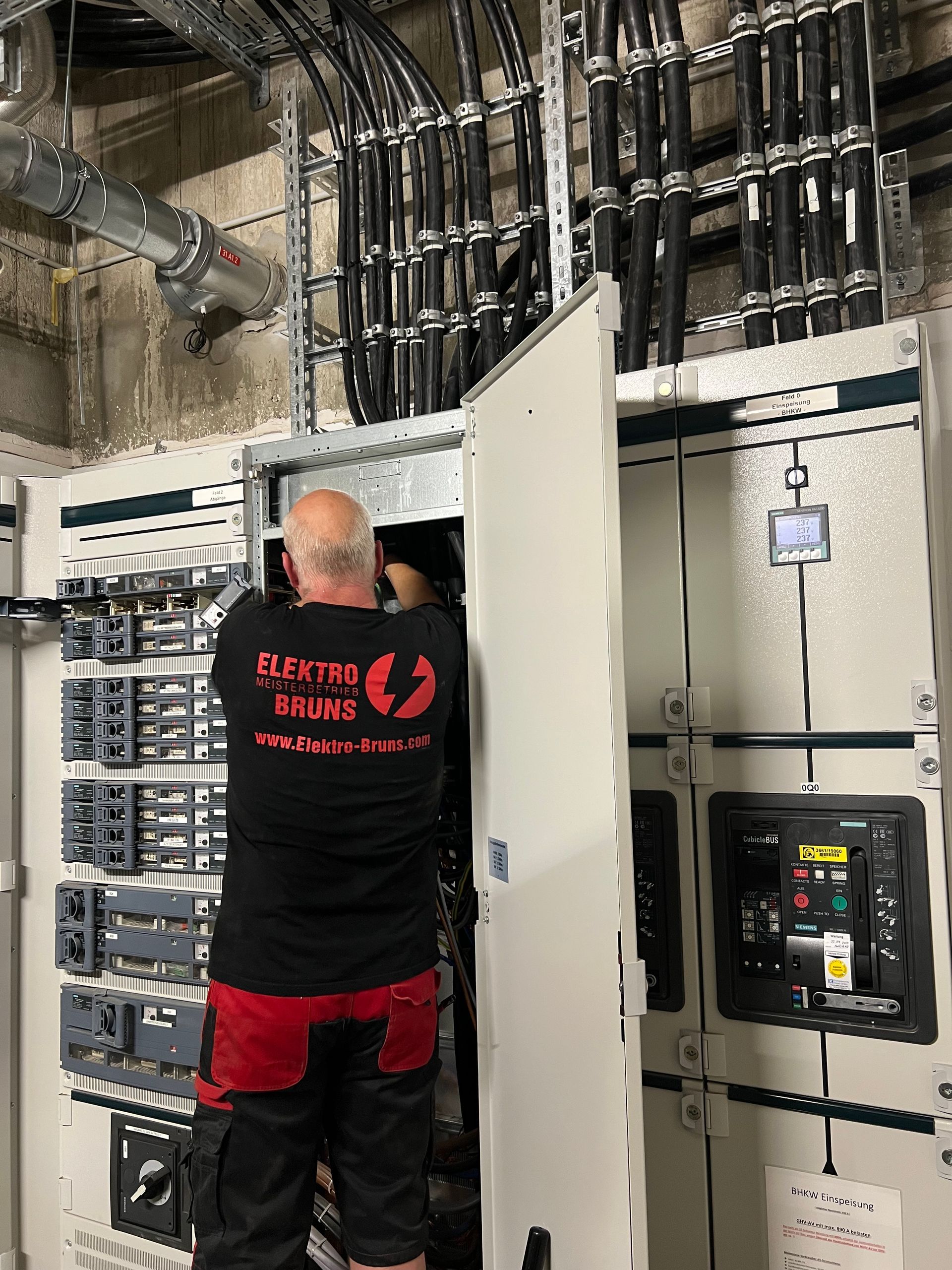 Techniker, der in einem elektrischen Kontrollraum mit offenen Schalttafeln und freiliegenden Leitungen arbeitet