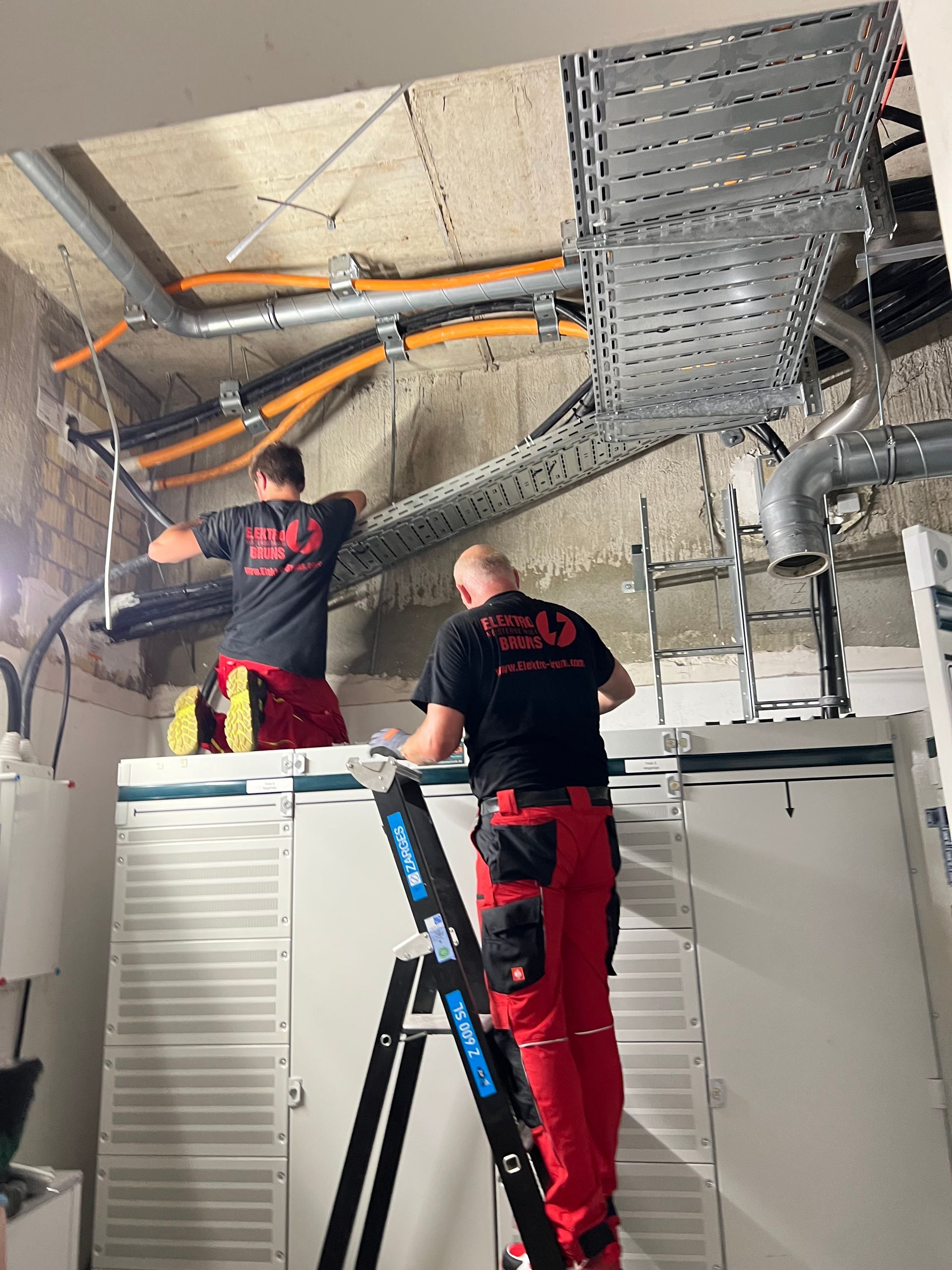 Zwei Arbeiter installieren auf Leitern Freileitungskabel in einem Technikraum mit freiliegenden Lüftungskanälen und Deckenplatten.
