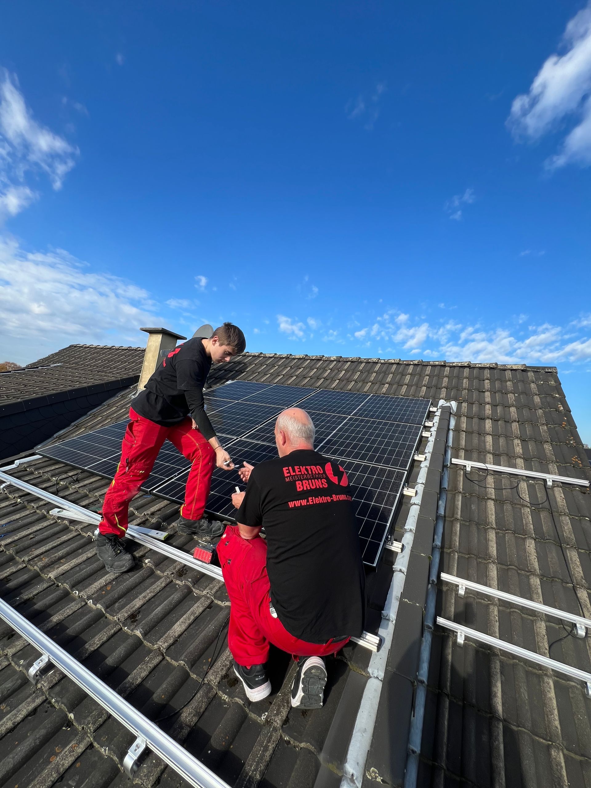 Zwei Arbeiter in roten Hosen auf einem steilen Dach, die unter blauem Himmel Solarpaneele inspizieren oder reparieren.