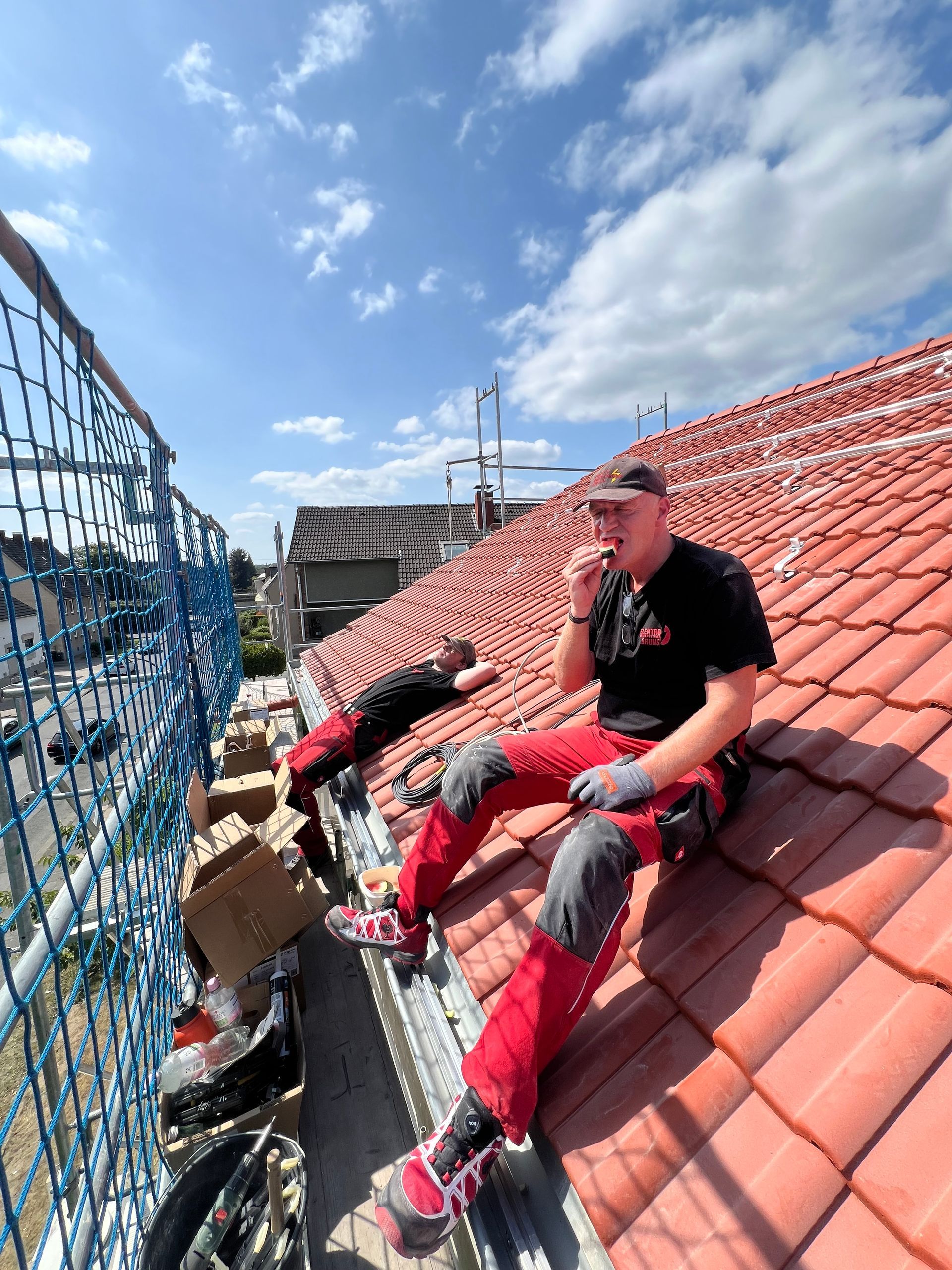 Ein Arbeiter sitzt an einem sonnigen Tag auf einem roten Ziegeldach neben einem Gerüst.