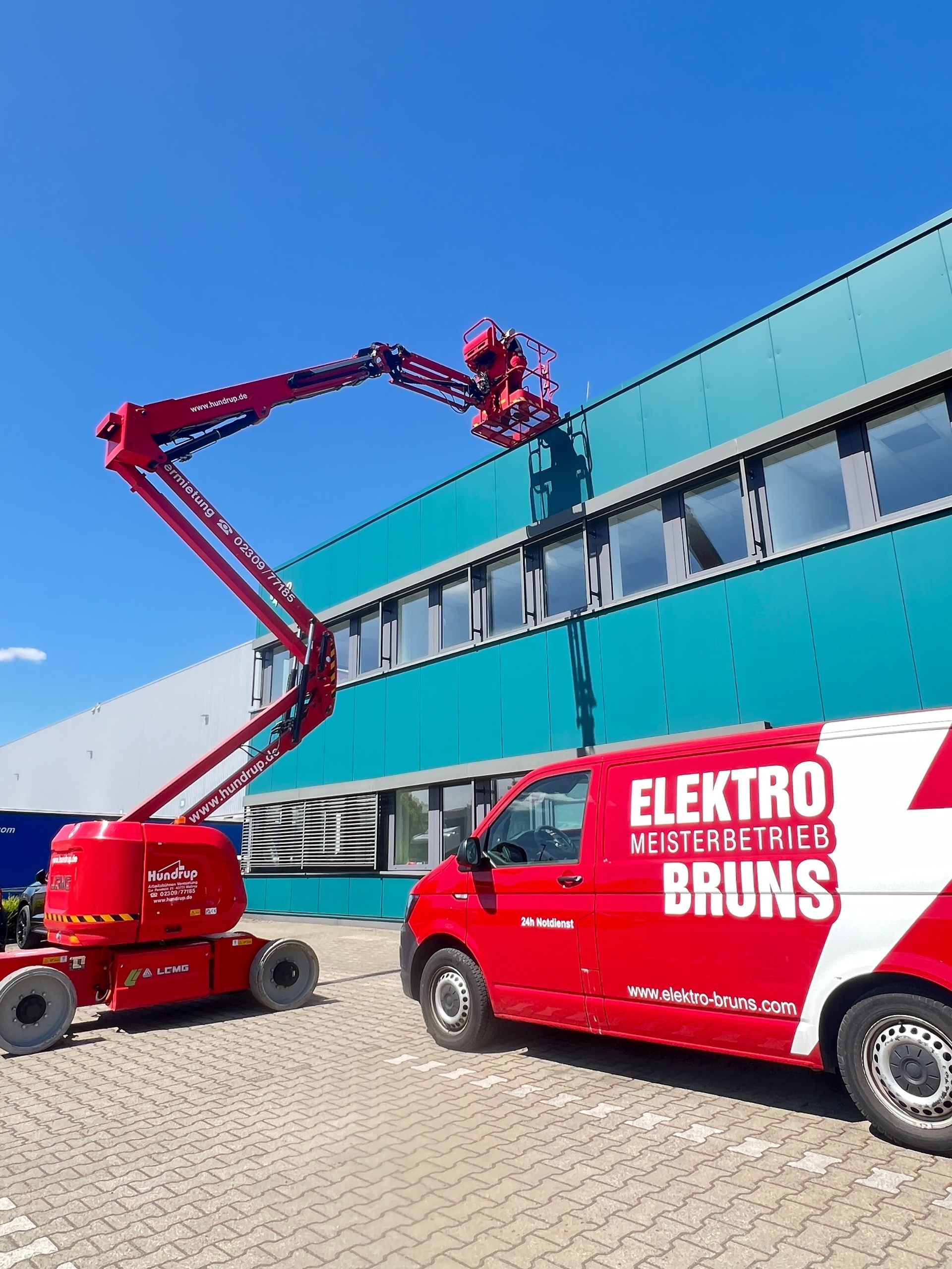 Ein roter Elektrogabelstapler mit Ausleger arbeitet neben einem türkisfarbenen Industriegebäude unter klarem, blauem Himmel.