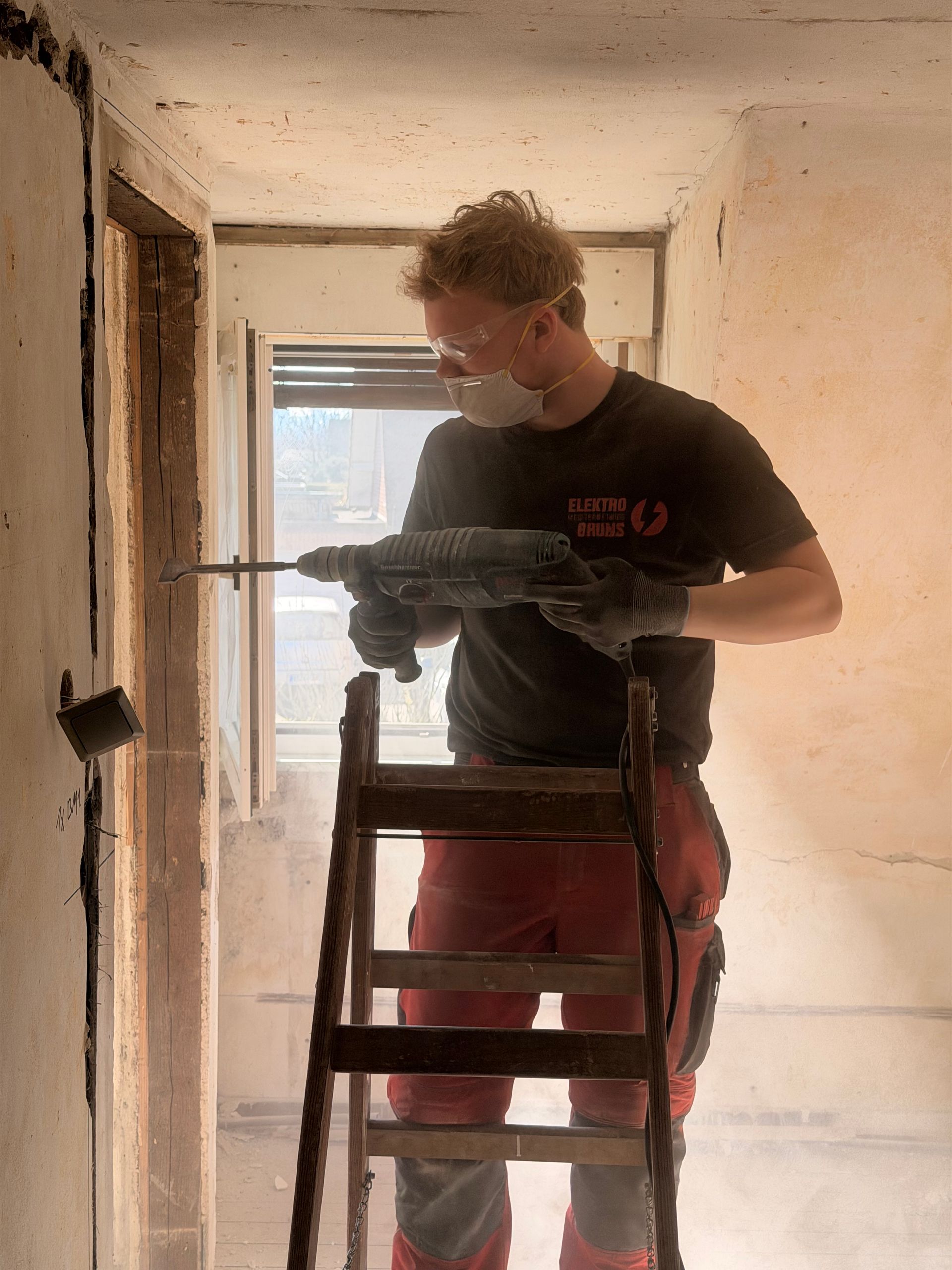 Ein Arbeiter steht auf einer Leiter und bohrt in eine Wand in einem schmalen, unfertigen Flur.