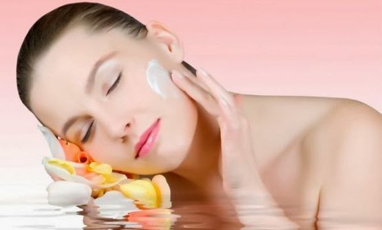 Mujer relajándose con crema blanca en la cara junto a flores sobre un fondo rosa suave.