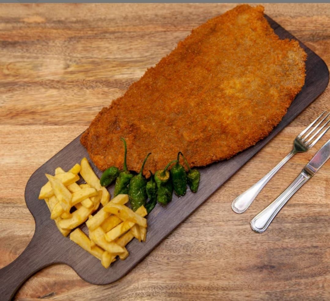 Un gran trozo de carne empanizada con patatas fritas y pimientos sobre una tabla de cortar de madera.