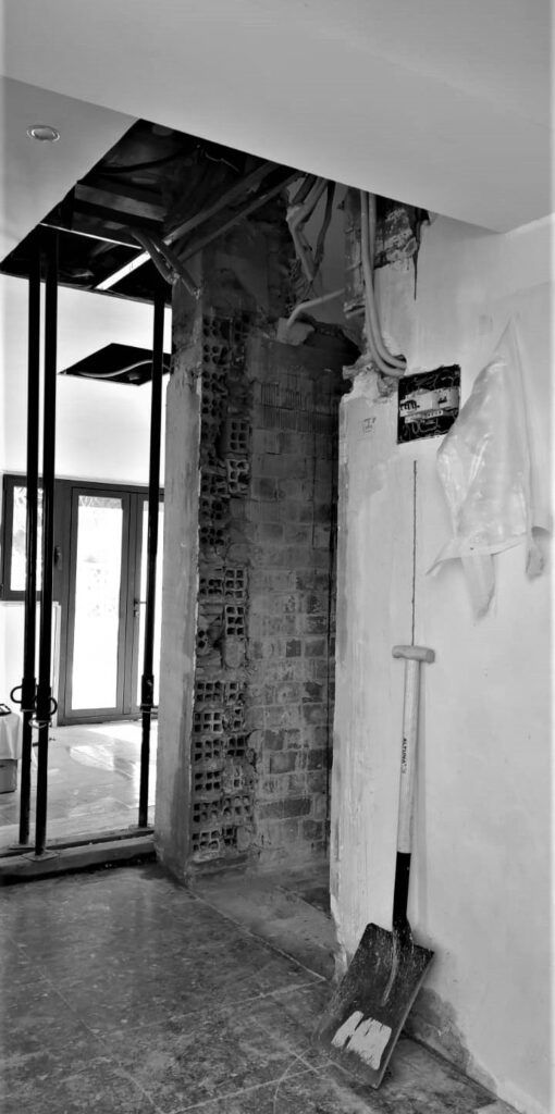 Escena de construcción interior con ladrillos a la vista, andamios y una pala apoyada contra una pared.