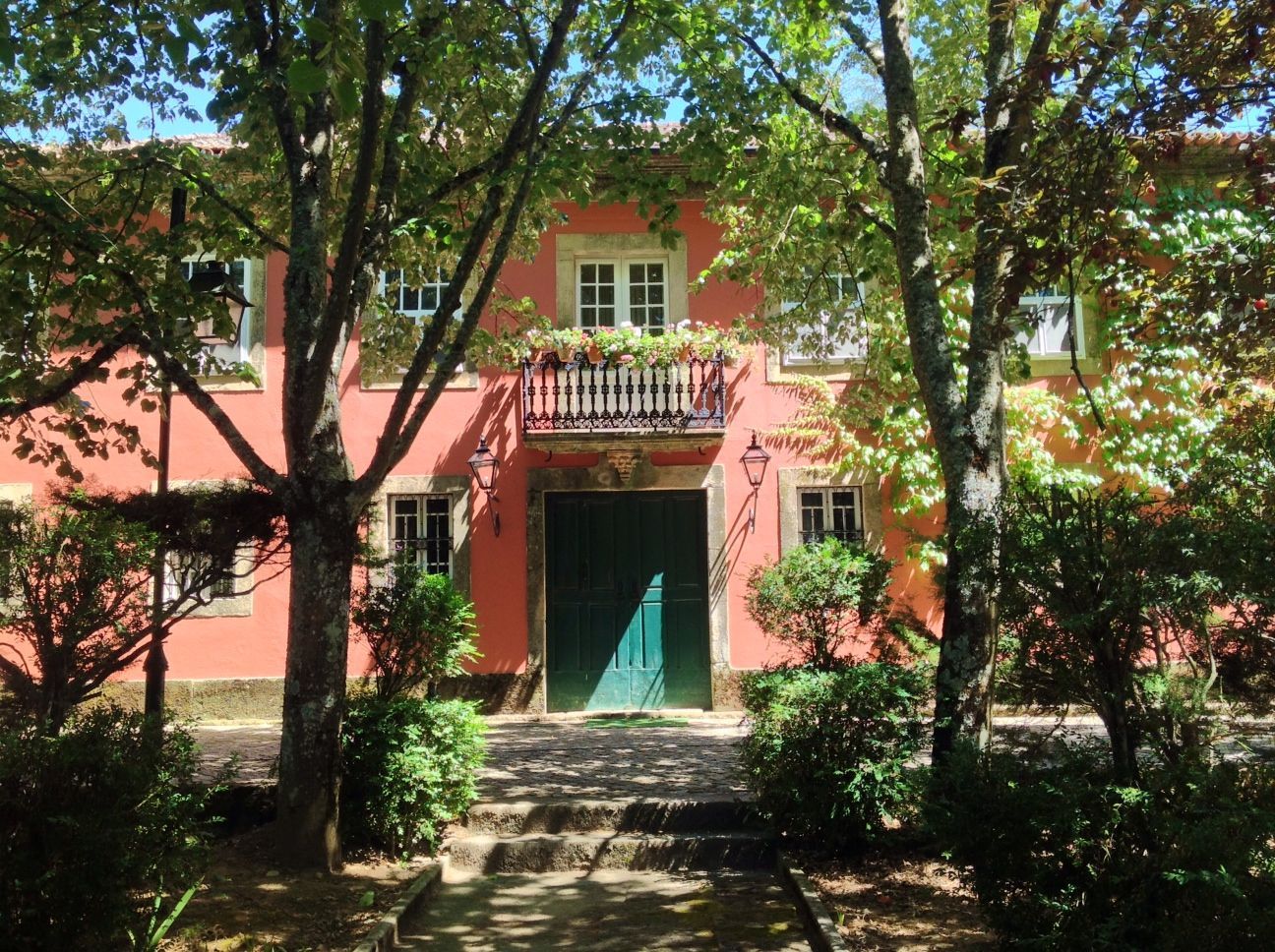 Uma casa rosa com uma porta verde cercada por árvores e arbustos.