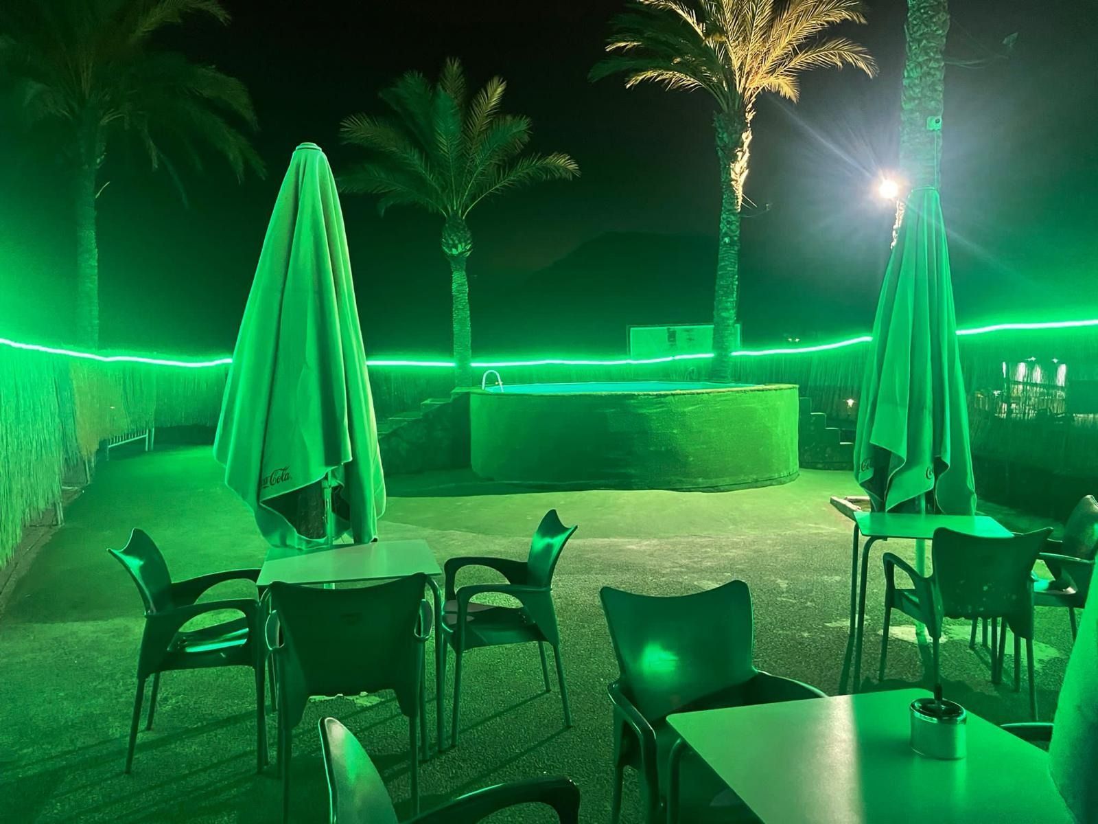 Un patio con mesas, sillas y sombrillas por la noche con luces verdes.