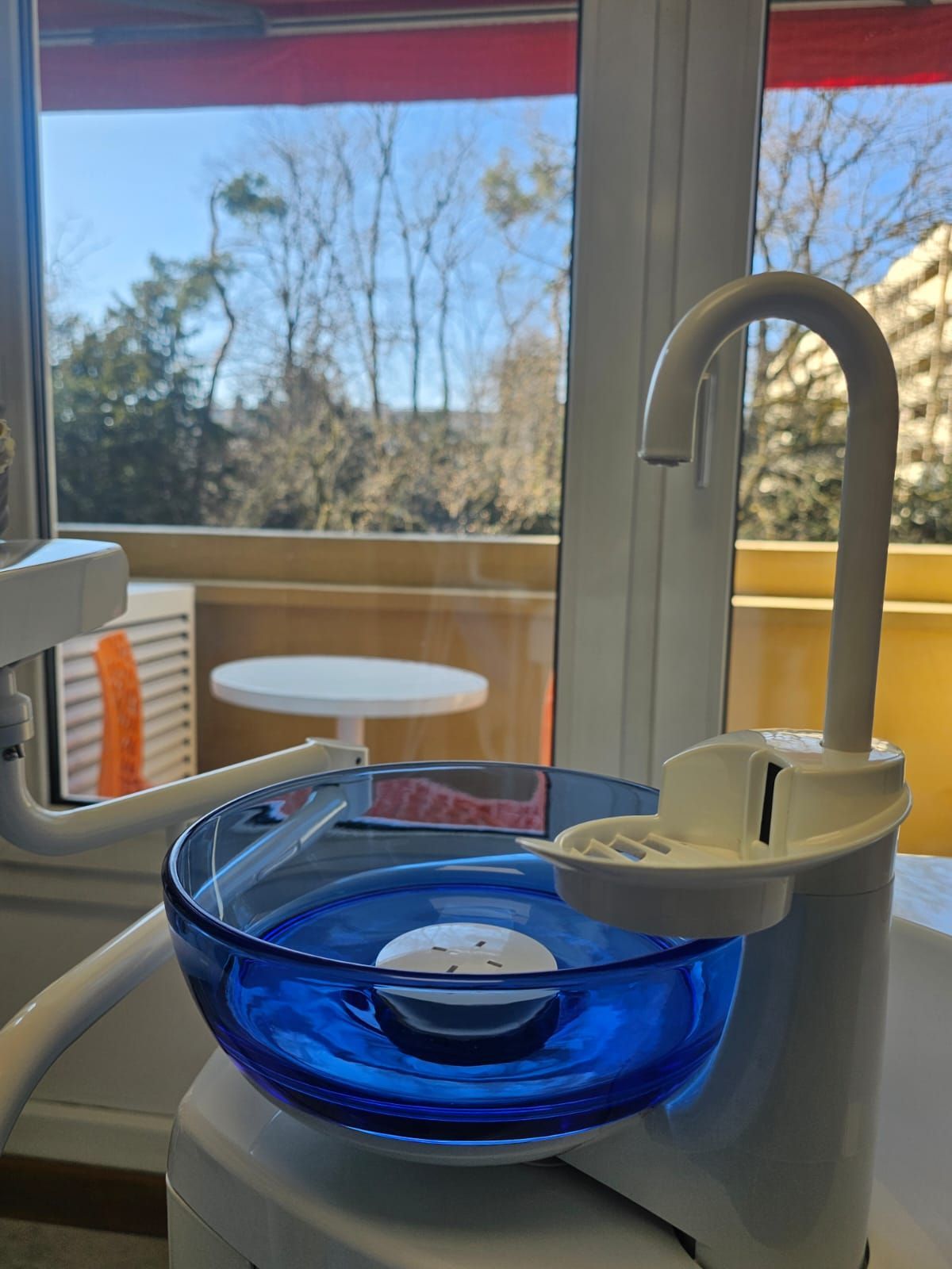 Un fauteuil dentaire avec un bol d'eau bleu devant une fenêtre.