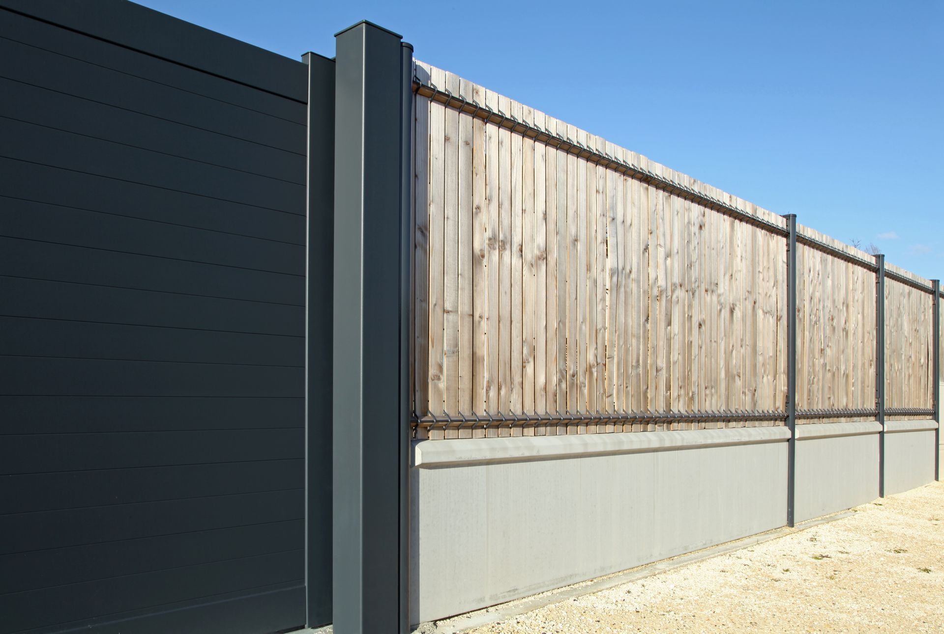 Haute clôture en bois avec des poteaux métalliques le long d'un mur en béton sous un ciel bleu clair