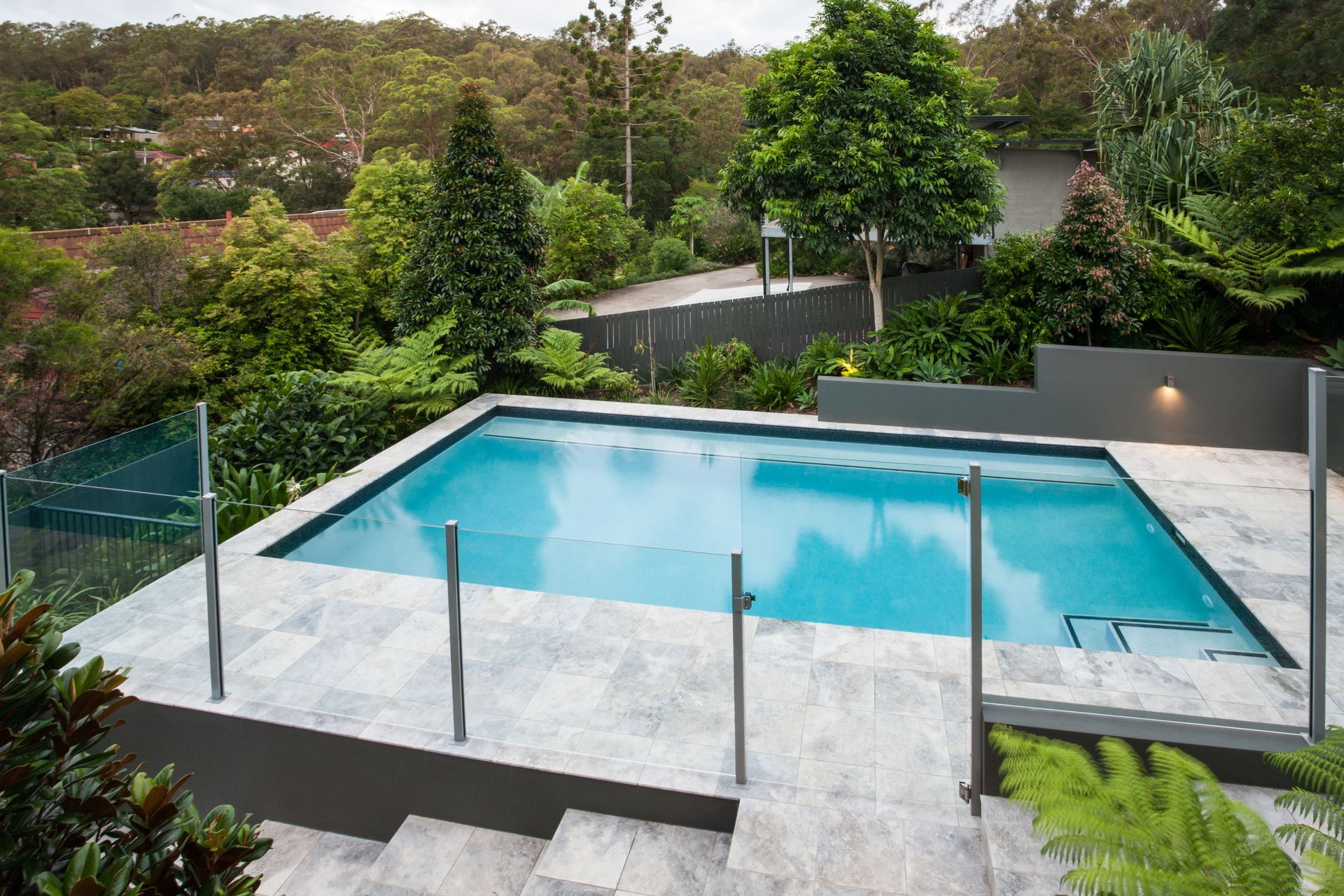 Piscine moderne dans le jardin avec clôture en verre, terrasse grise et végétation luxuriante au-delà