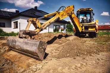 Une pelleteuse jaune travaille dans un terrain vague à côté d'une maison, déplaçant de la terre avec un godet.