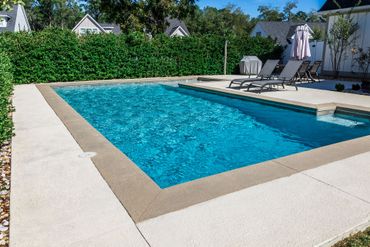 Piscine rectangulaire turquoise avec margelles beiges, terrasse blanche et chaises longues dans un jardin ensoleillé