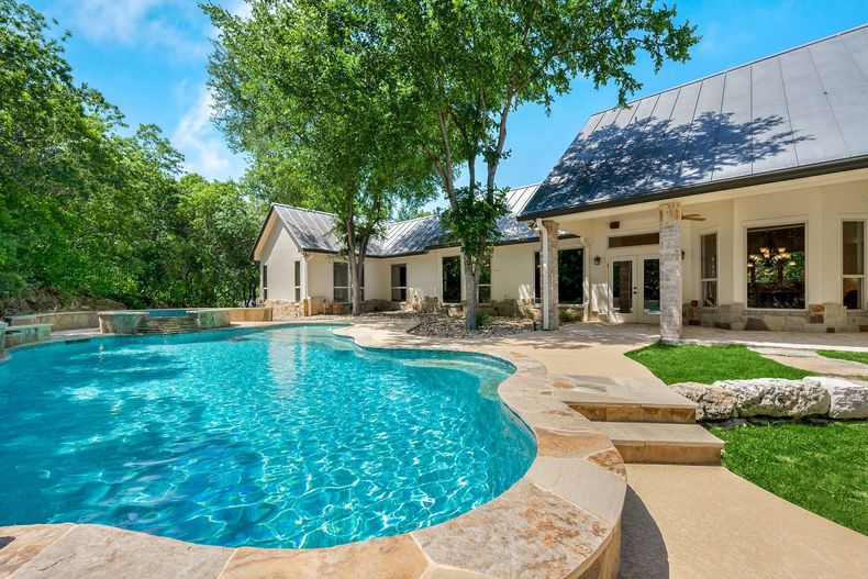 Piscine dans le jardin avec terrasse en pierre, grands arbres et patio d'une maison blanche sous un ciel bleu