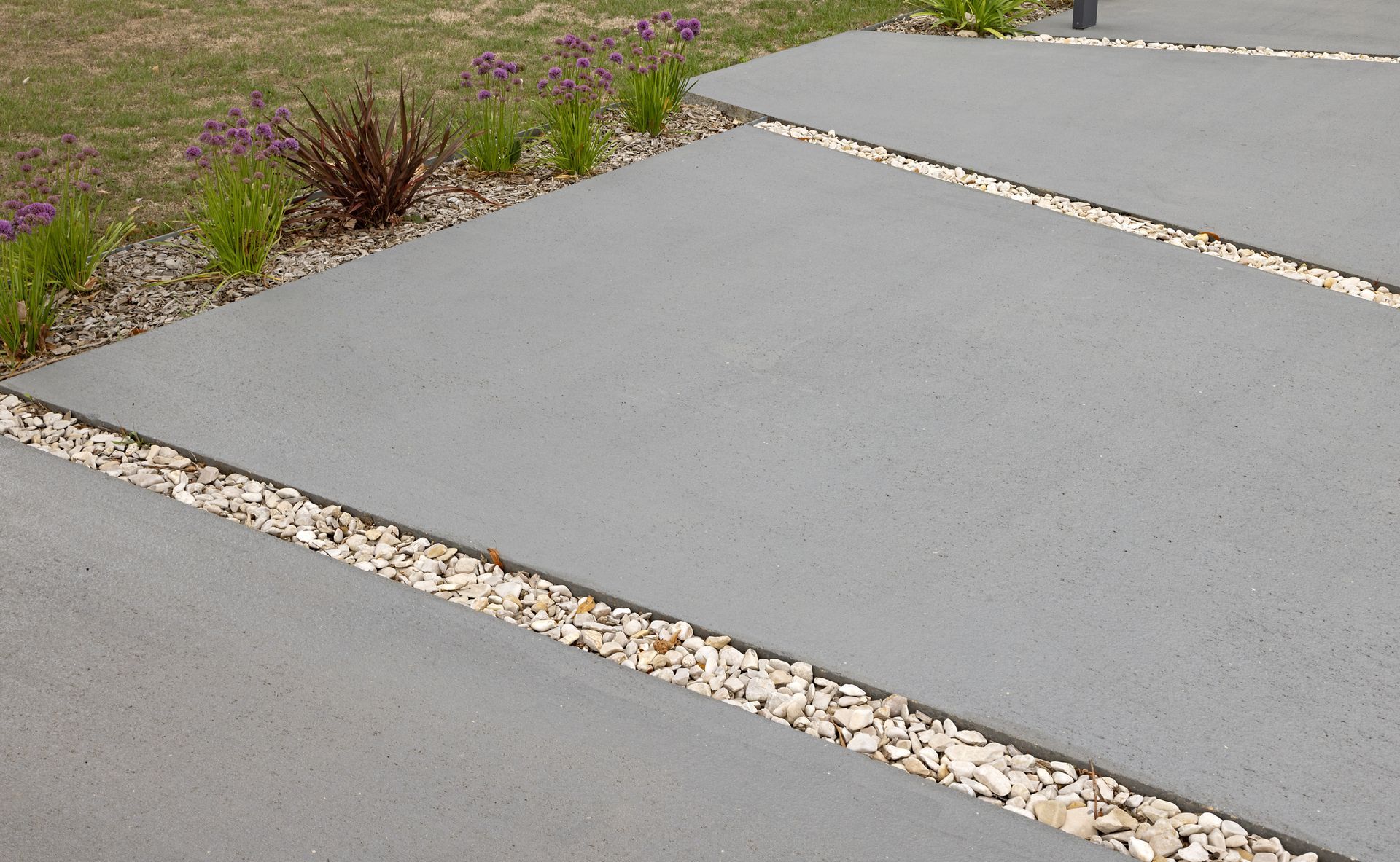 Allée en béton gris avec joints de dilatation, bordée d'une pelouse recouverte de galets et de petites plantes.
