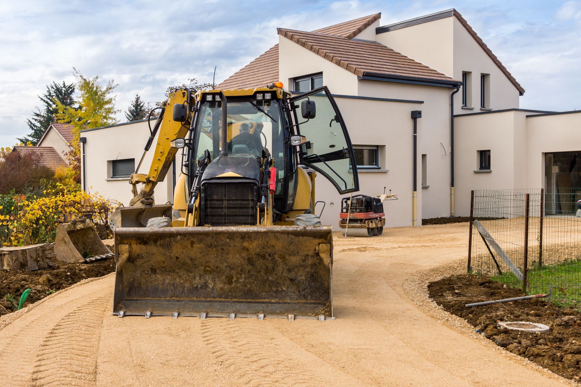 Une chargeuse-pelleteuse sur le chantier d'une maison d'habitation en construction, avec une allée en terre et des murs extérieurs blancs.