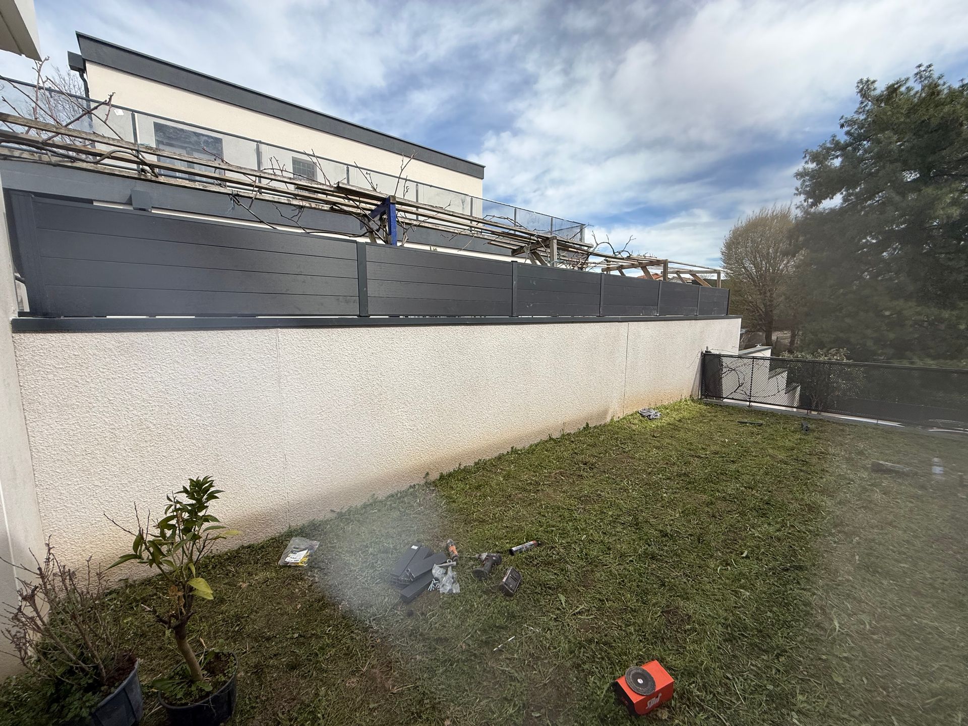 Cour herbeuse jouxtant un mur blanc et un bâtiment moderne, avec une petite voiture rouge au premier plan et un ciel nuageux au-dessus.