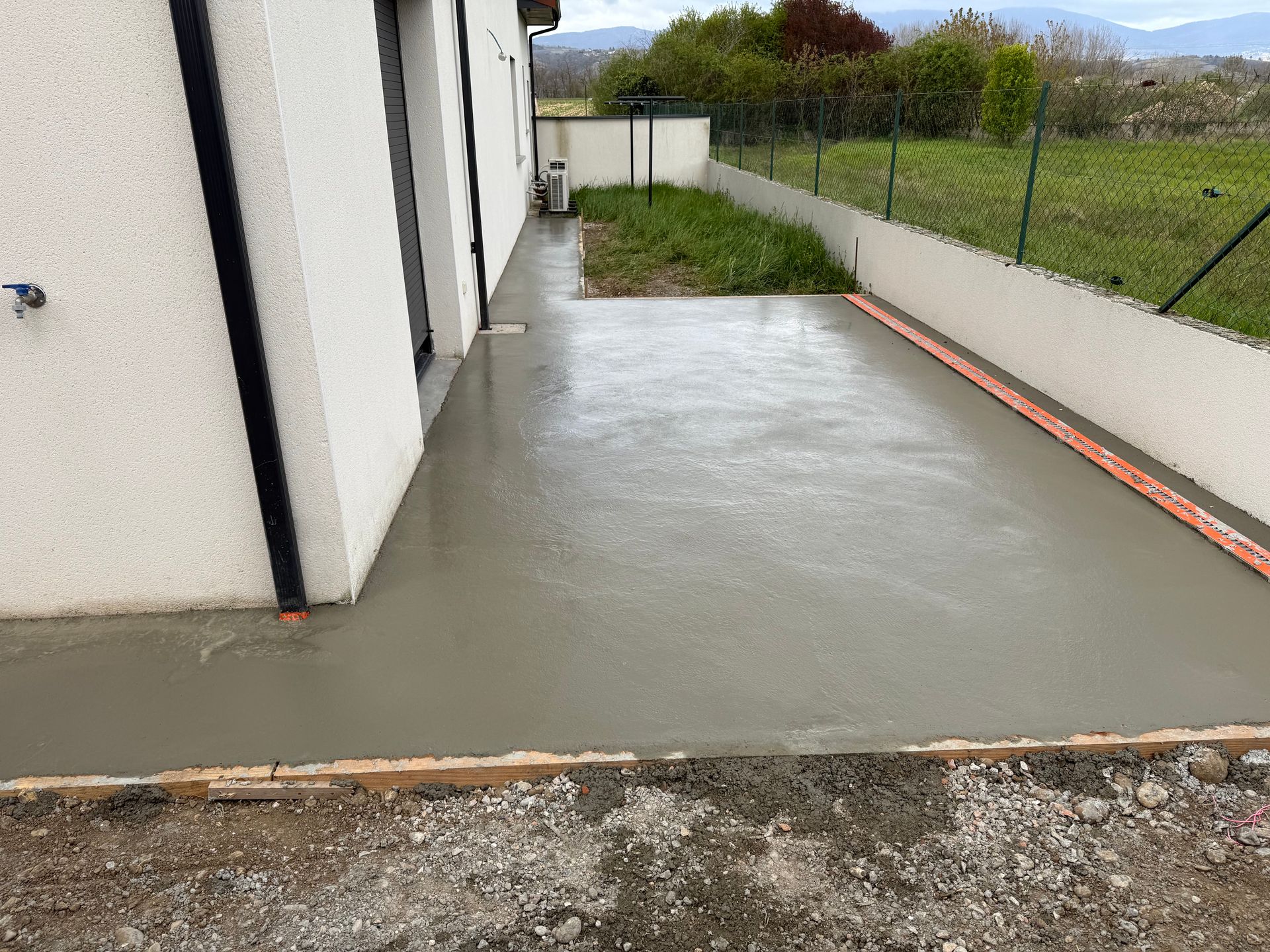 Terrasse en béton jouxtant un bâtiment blanc, mouillée par la pluie, avec une cour gazonnée clôturée en arrière-plan.