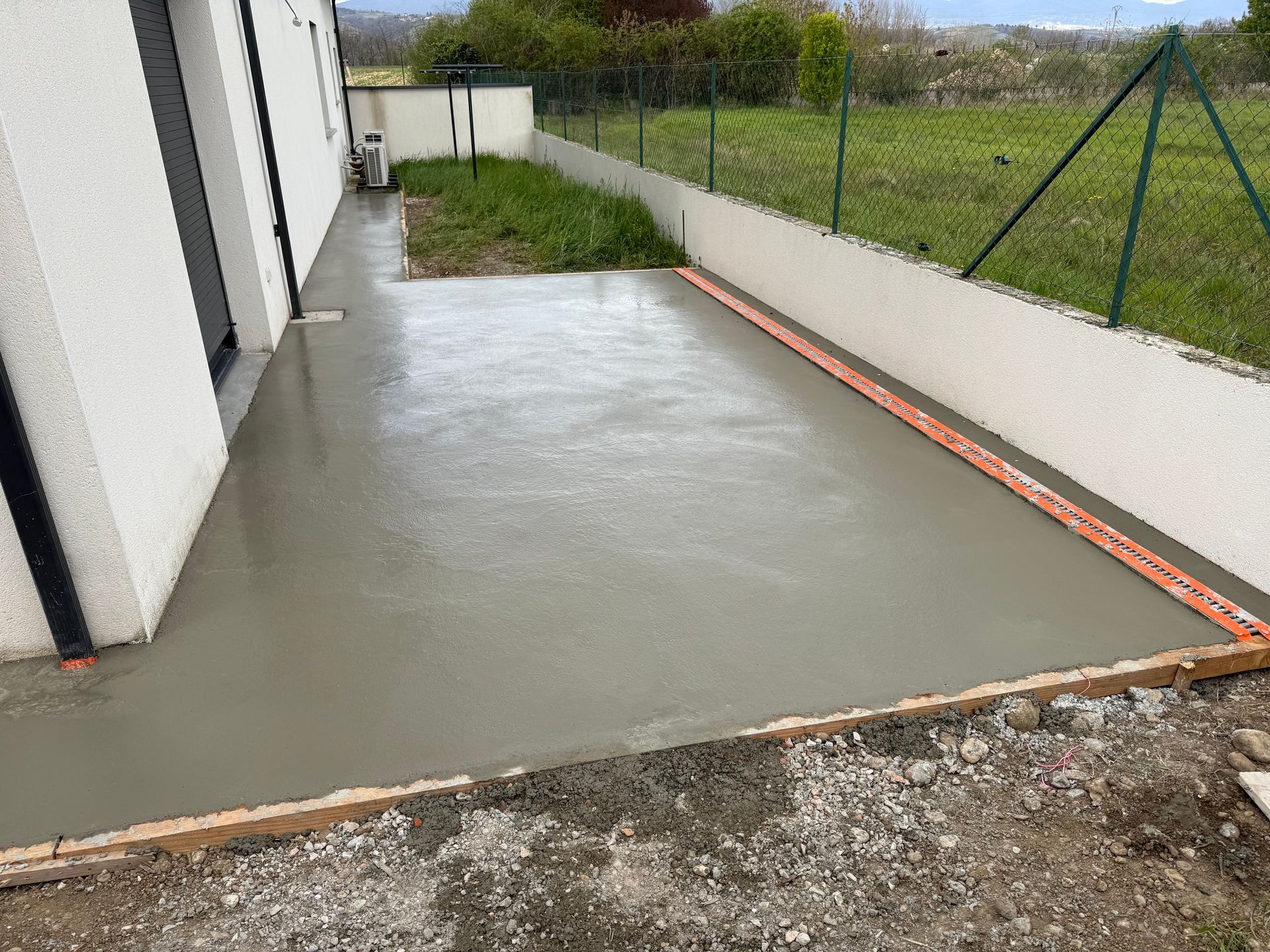 Terrasse en béton neuf jouxtant une maison blanche, bordée de gravier et donnant sur une cour gazonnée clôturée.