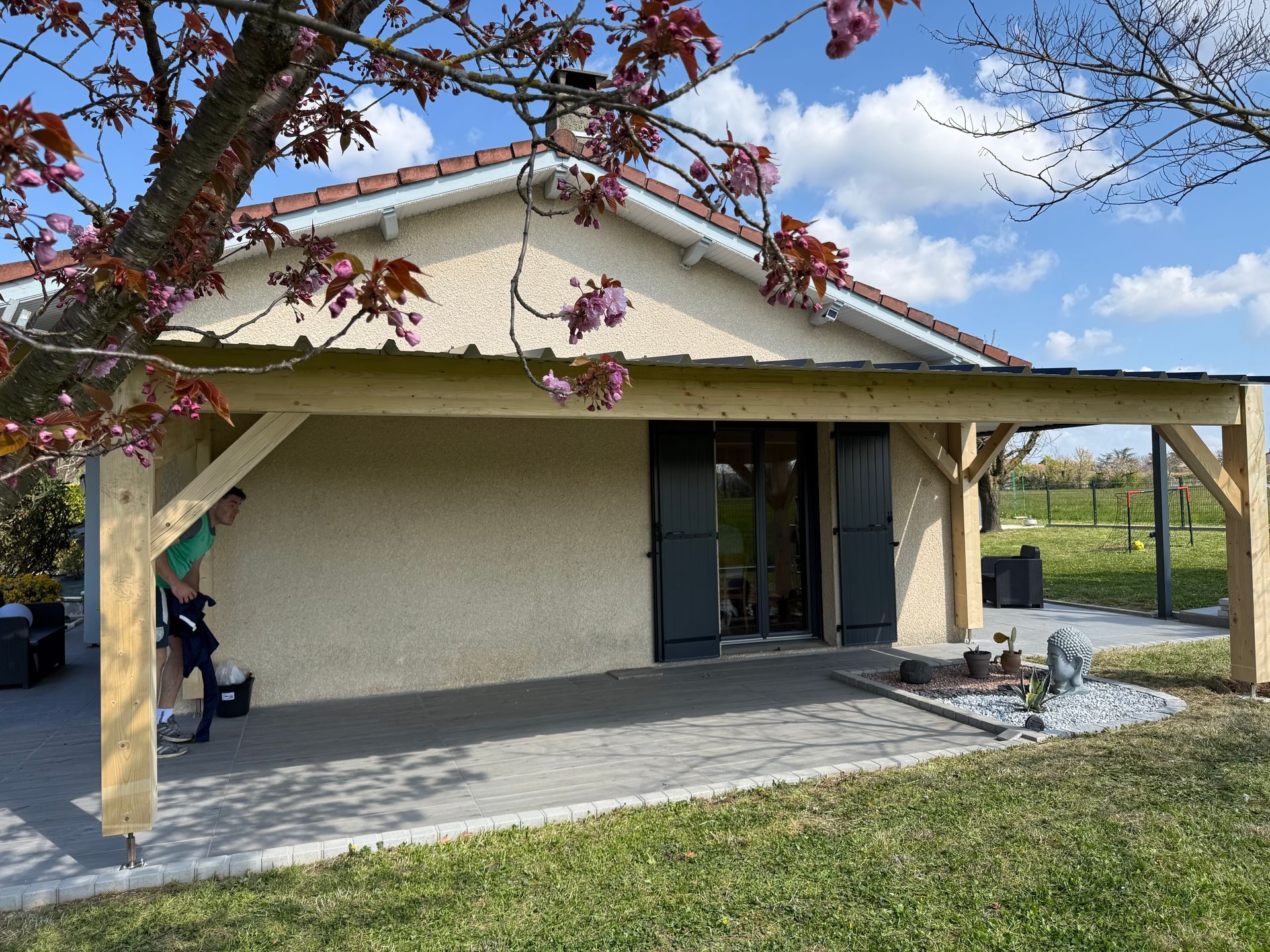 Extérieur de maison avec patio couvert, murs en stuc beige et une branche d'arbre au-dessus par une journée ensoleillée