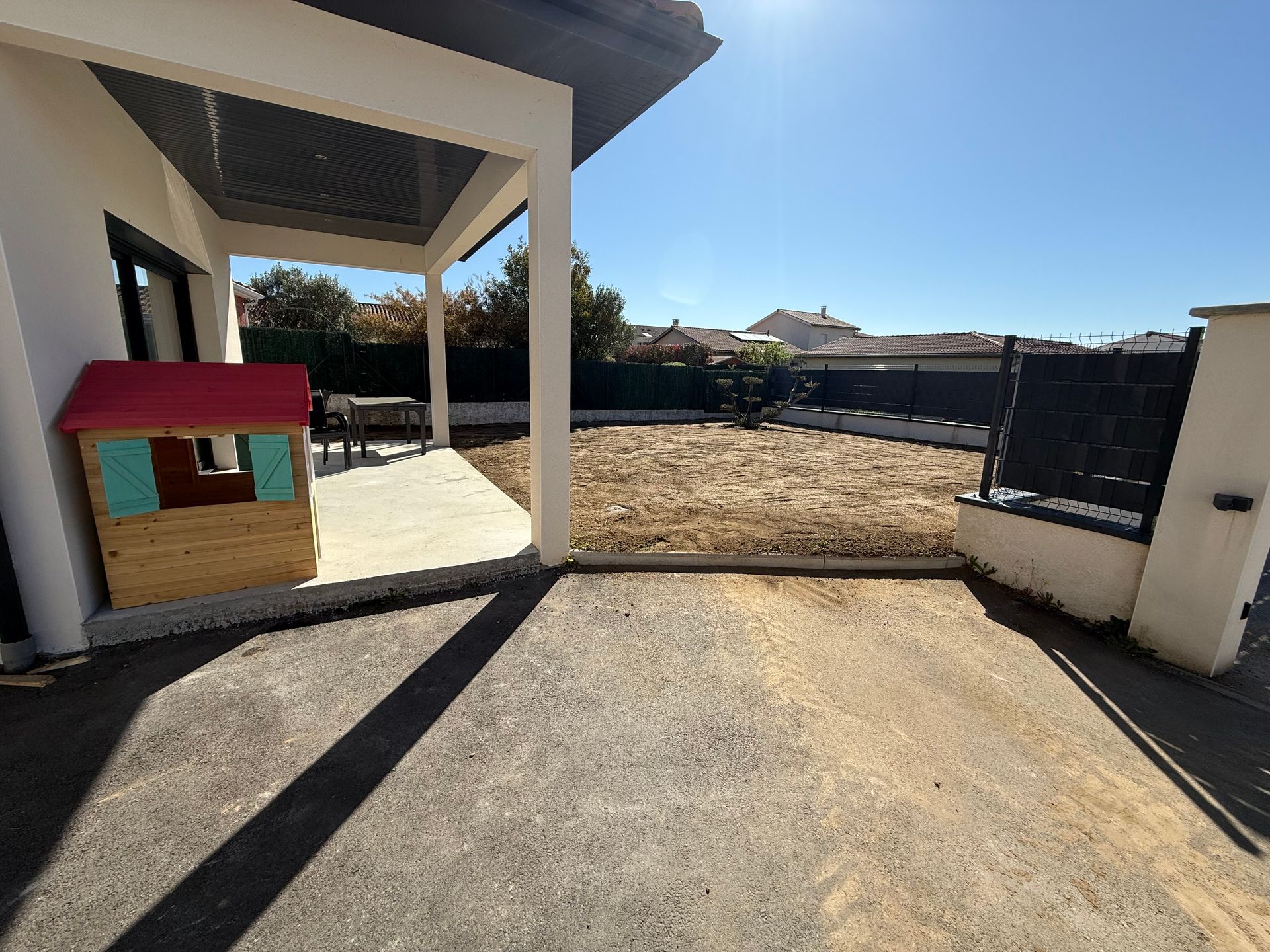 Allée ensoleillée longeant une terrasse couverte, avec une petite maisonnette rouge et verte sur la gauche.