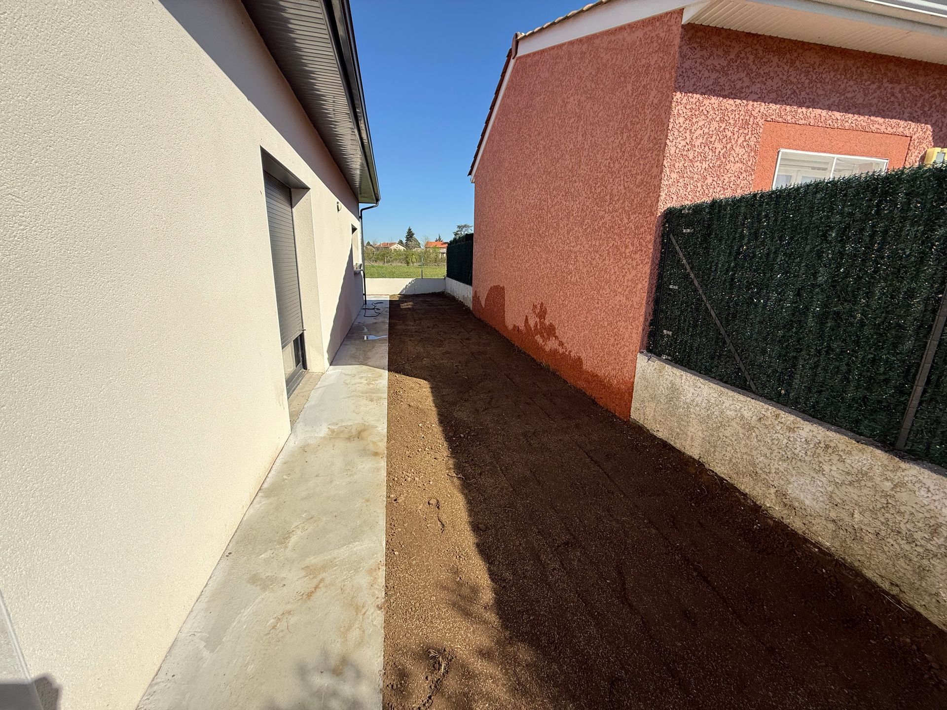 Passage extérieur étroit entre une maison beige et un mur rouge, avec un sol en gravier et des haies sur la droite.
