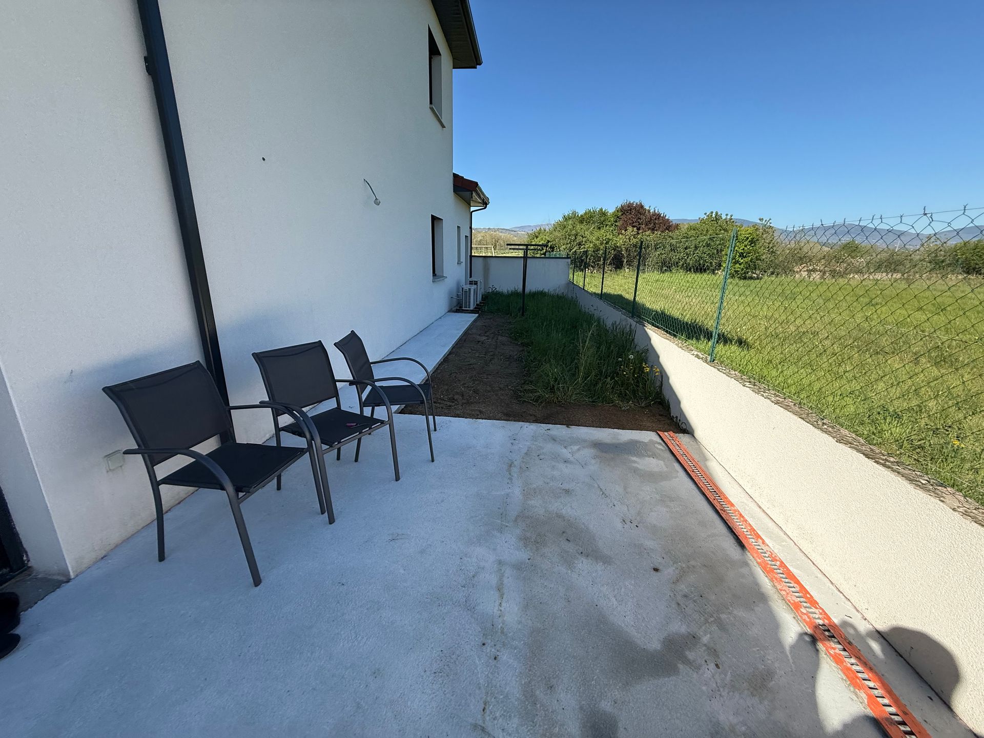 Terrasse extérieure jouxtant une maison blanche, avec chaises noires, sol en béton et jardin gazonné au-delà d'un muret.