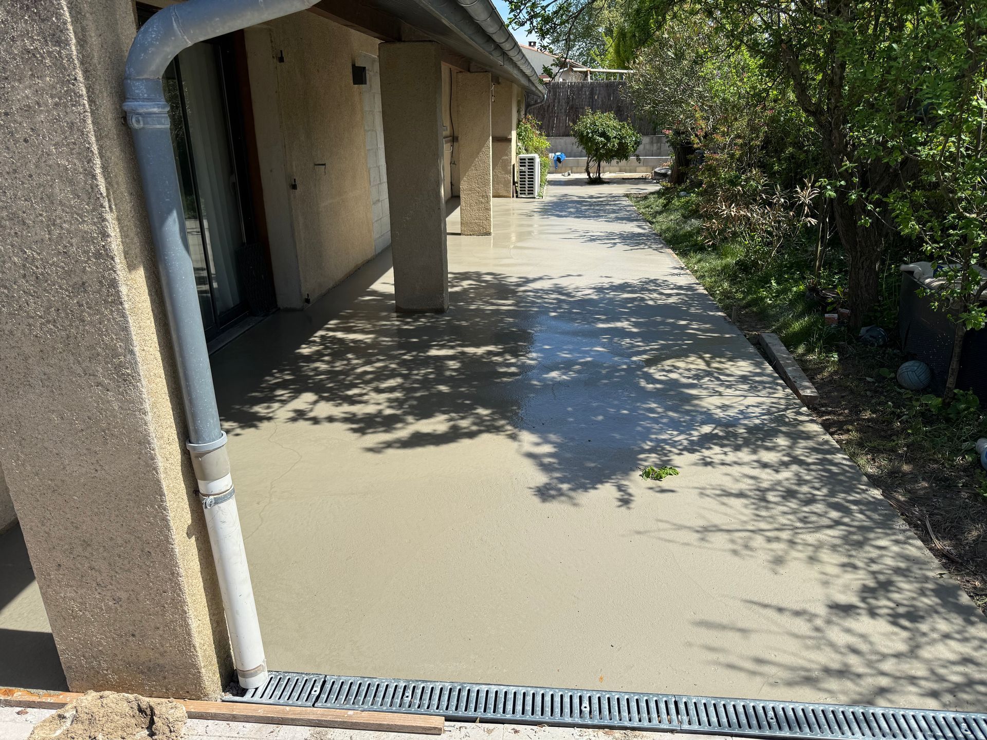 Allée en béton longeant un bâtiment, avec une entrée ombragée et des arbres bordant le chemin.