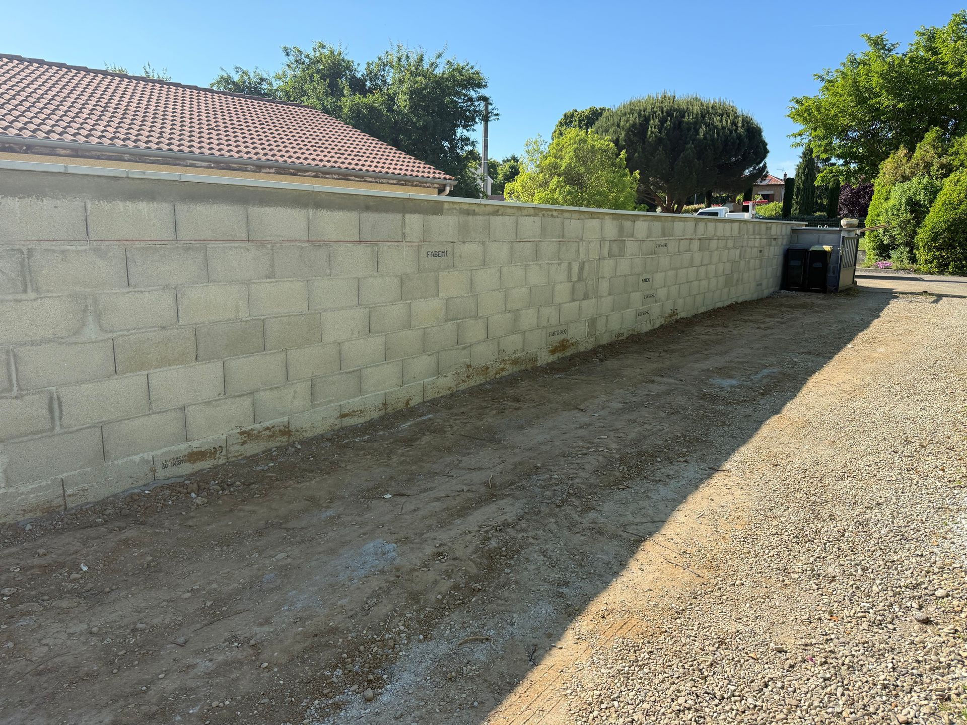 Allée de gravier longeant un long mur de parpaings et un toit de tuiles, avec des arbres et un portail au loin.