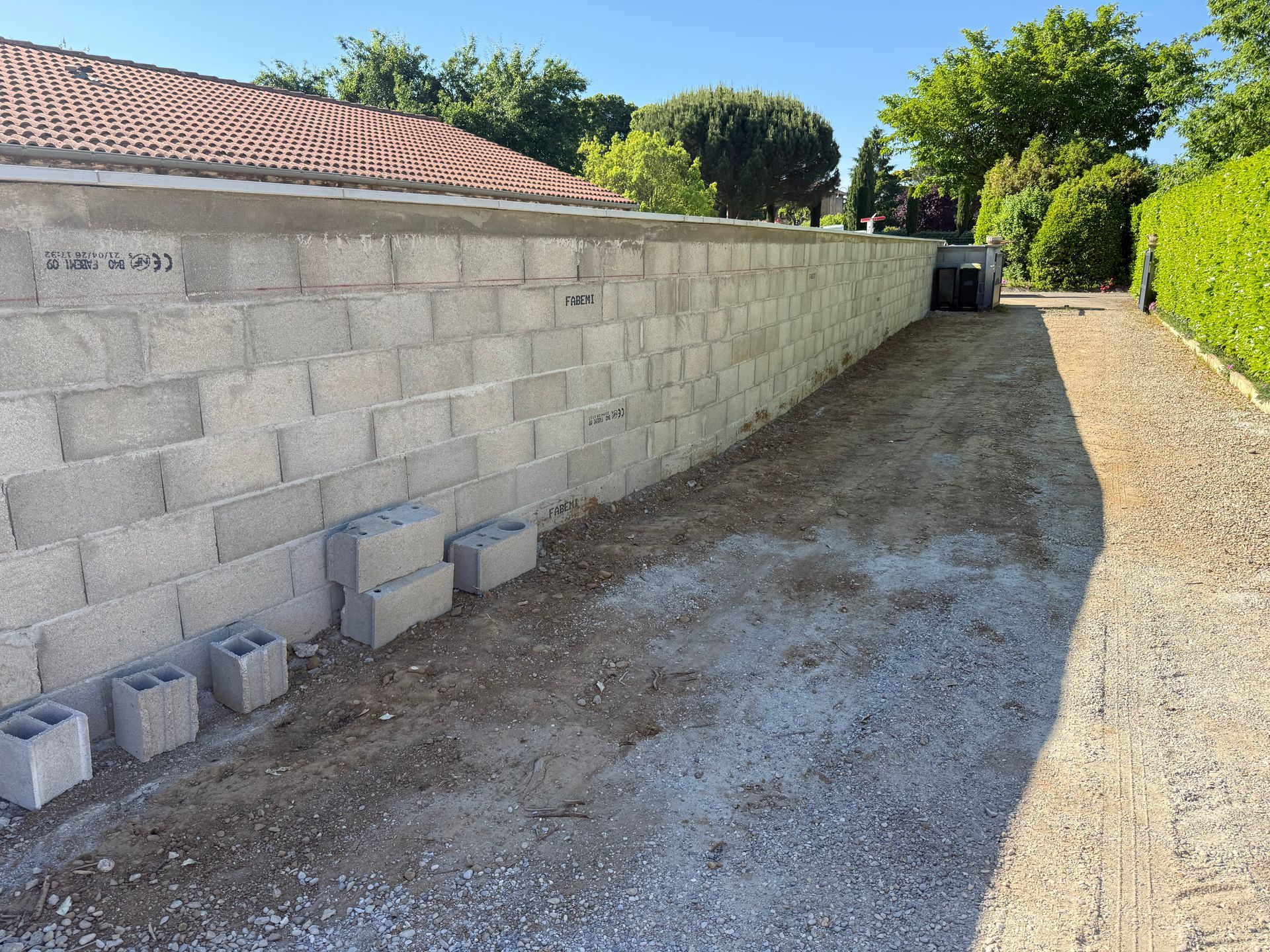 Long mur en parpaings bordant une allée de gravier, baigné de soleil, avec des arbres et des buissons à proximité.