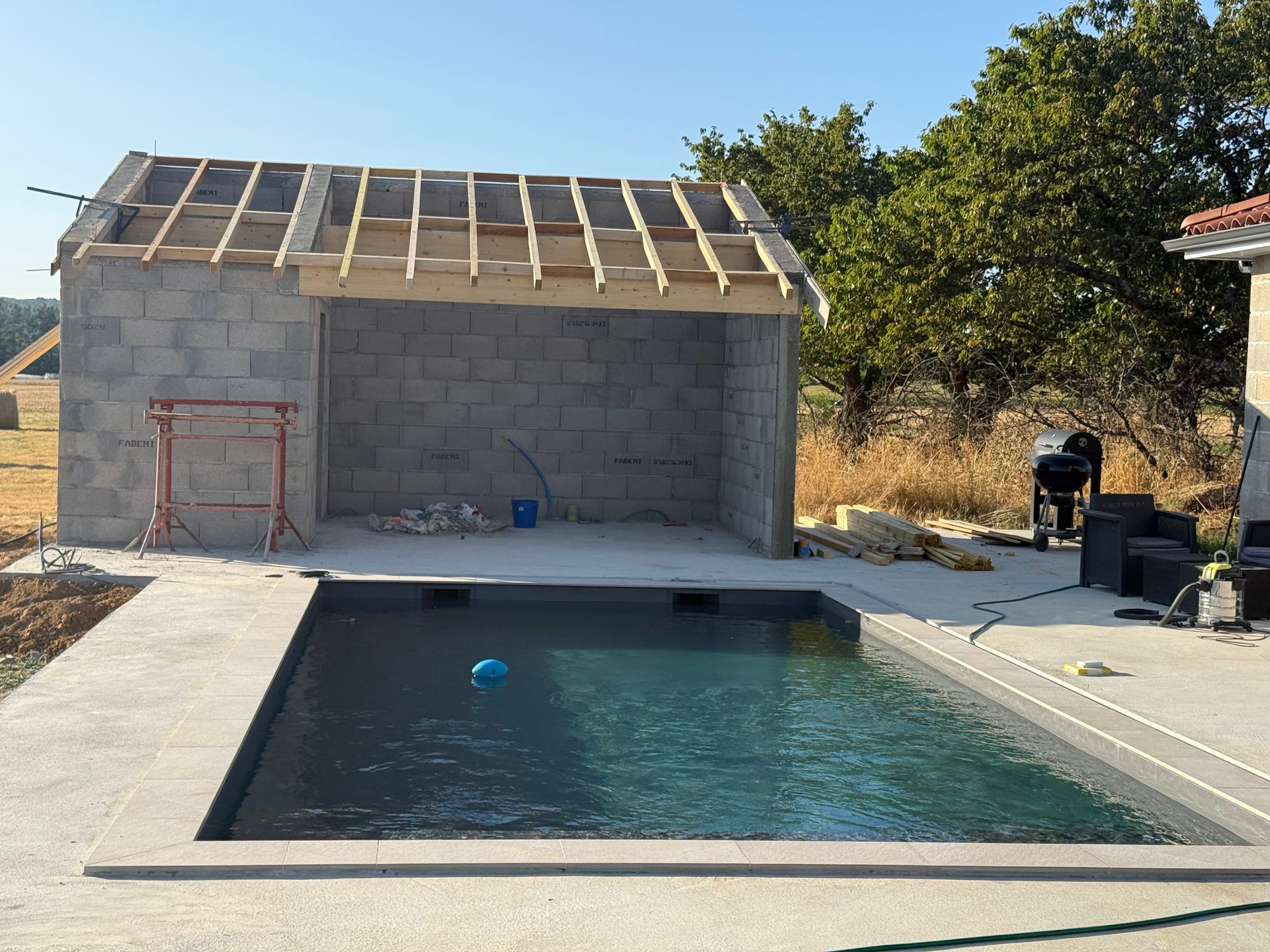 Piscine dans le jardin, à côté d'un abri en parpaings en construction, avec la charpente du toit et des arbres en arrière-plan.