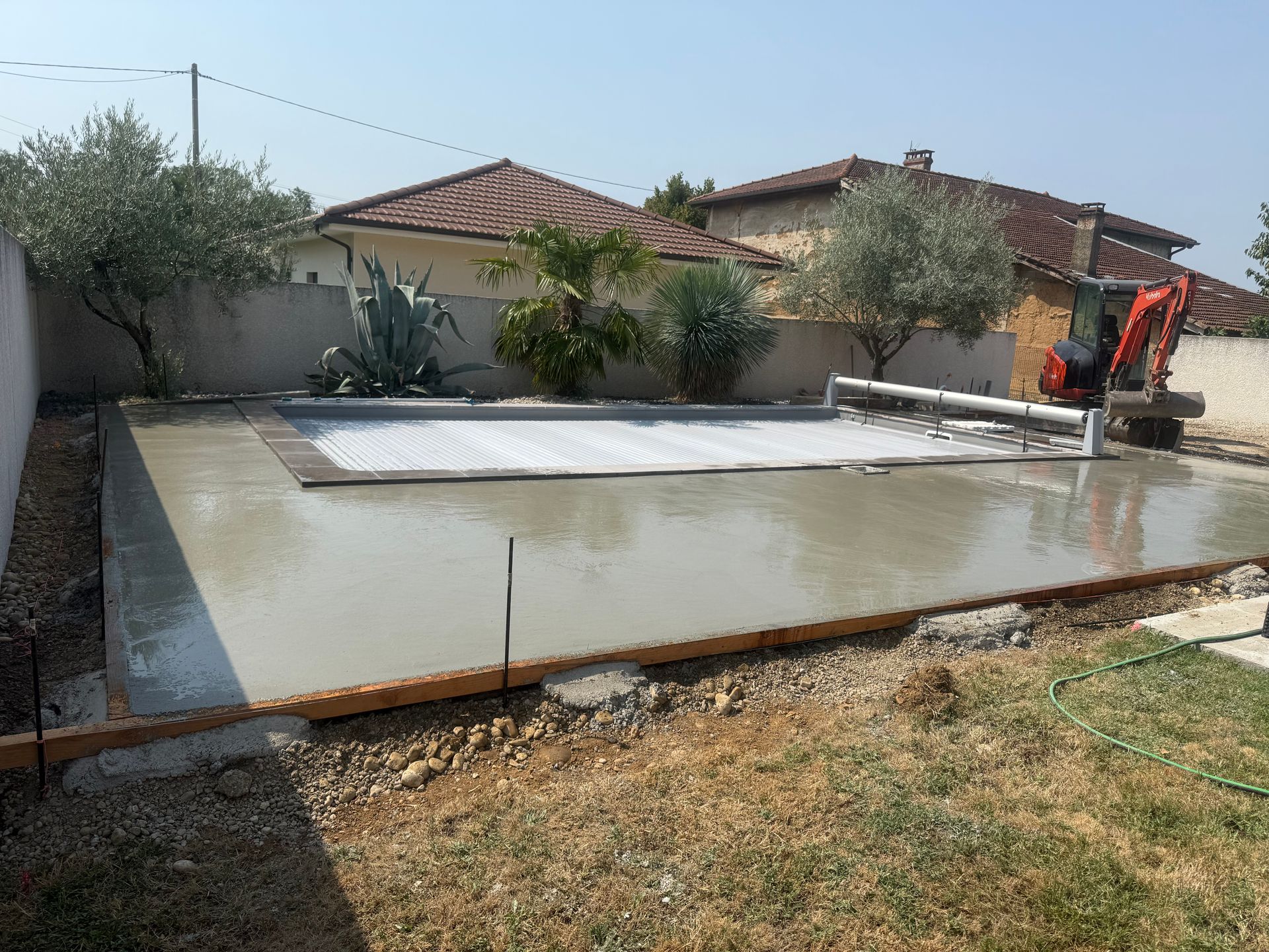 Dalle de béton fraîchement coulée dans une cour arrière, avec un engin de chantier et les maisons environnantes