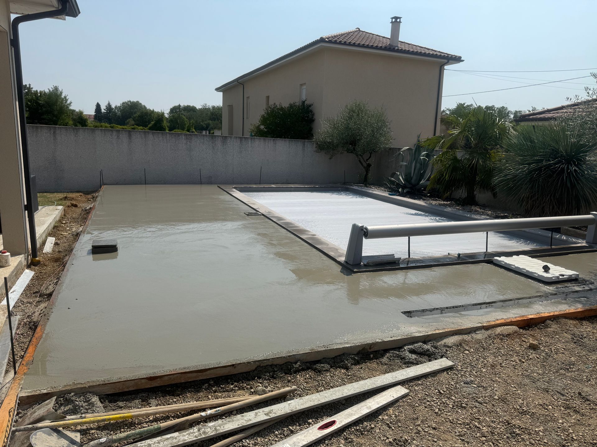 Dalle de béton partiellement coulée à côté d'une piscine couverte, avec du gravier et une maison en arrière-plan.