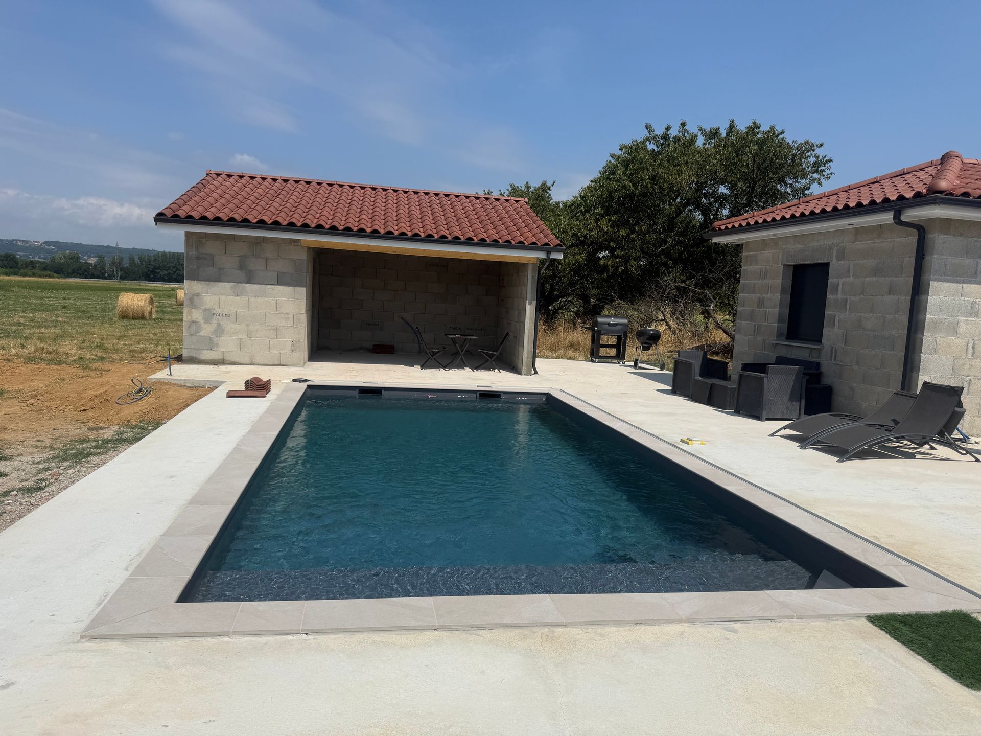 Piscine rectangulaire dans le jardin, jouxtant deux petits bâtiments en pierre aux toits de tuiles rouges.