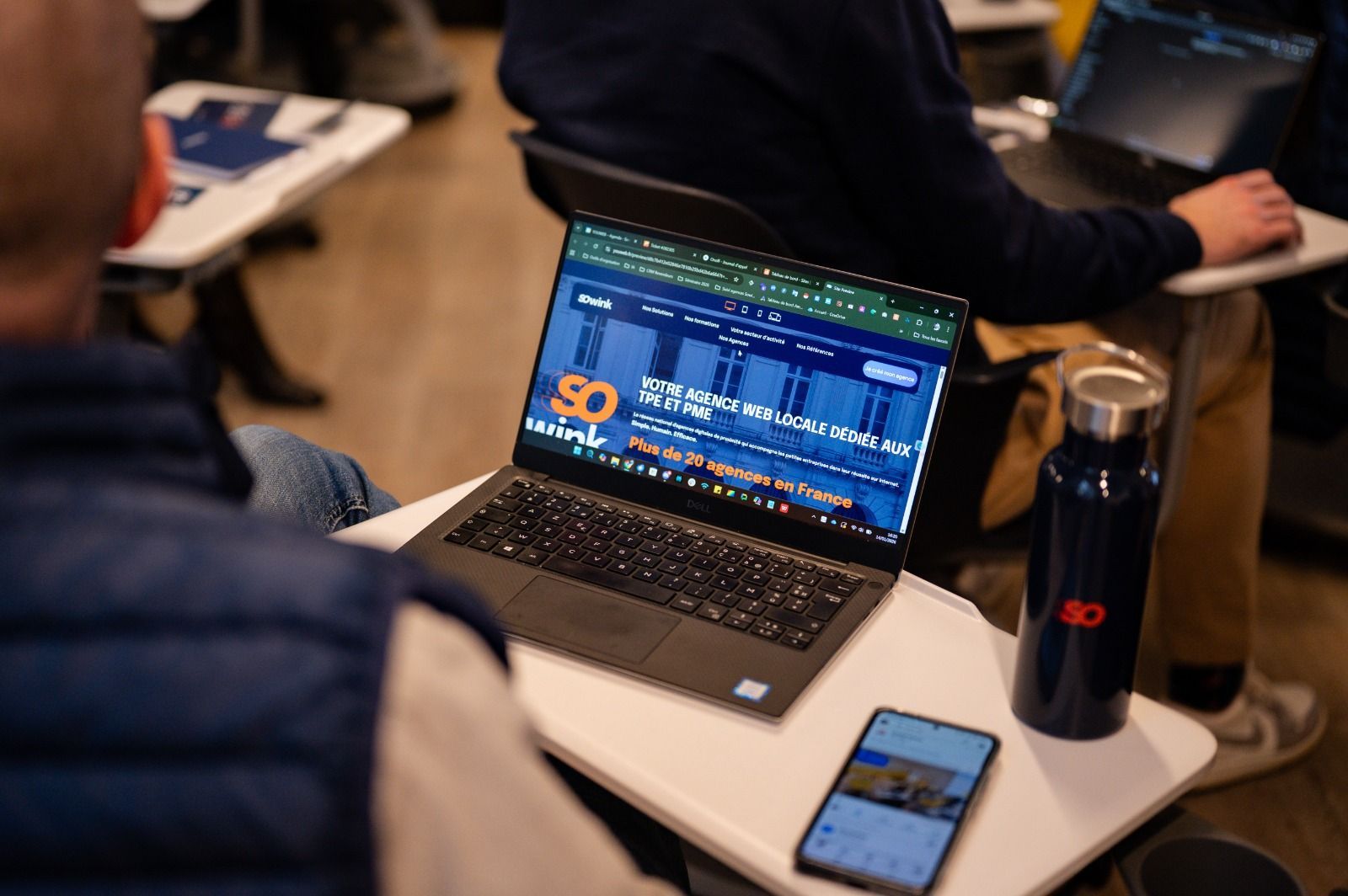 Un ordinateur portable posé sur un bureau affiche le site web Sowink, avec une bouteille d'eau et un téléphone assortis, dans un décor de salle de classe.