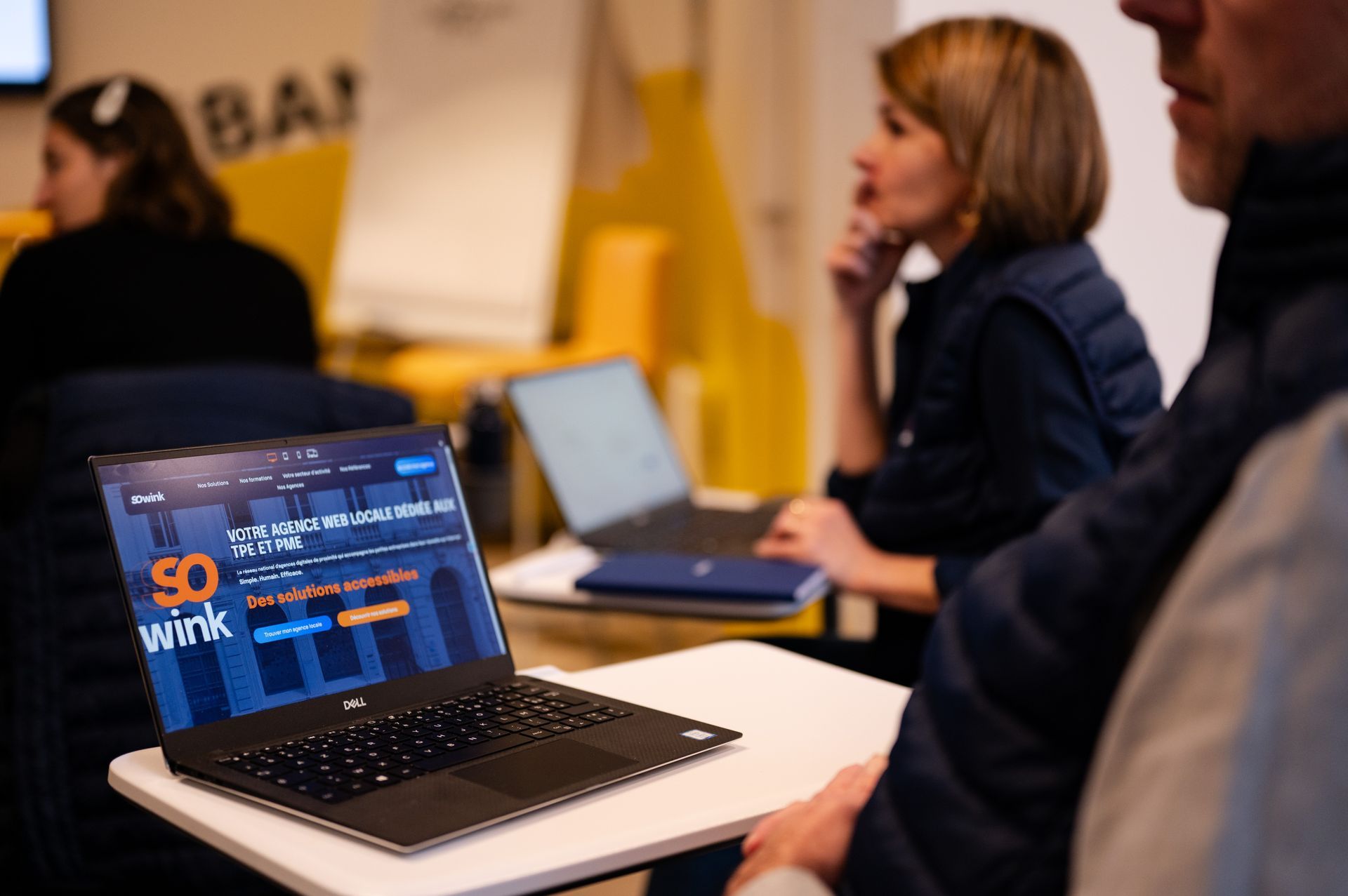 Un ordinateur portable affichant le site web « SOWINK » est posé sur un bureau dans une salle de classe, tandis que des personnes travaillent en arrière-plan.