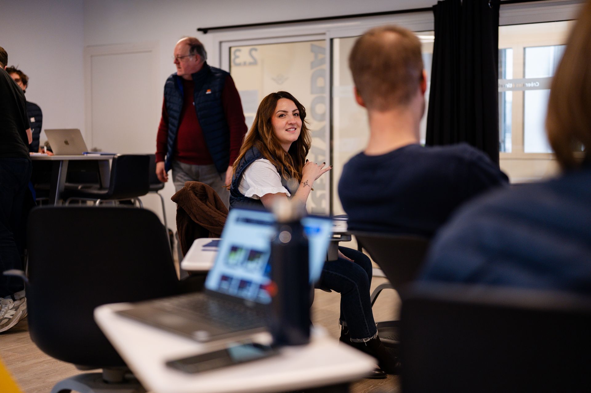 Des personnes sont assises dans un bureau lumineux ; une femme se tourne vers la caméra tandis qu'une autre personne travaille sur un ordinateur portable.