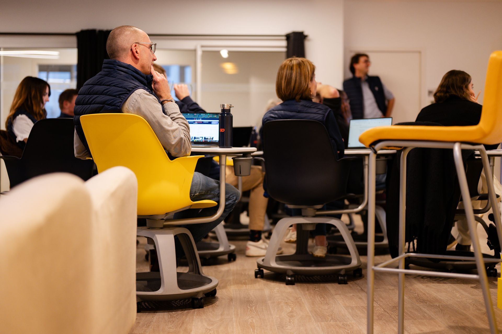 Dans un espace de bureaux moderne, des personnes sont assises à des tables avec des ordinateurs portables, concentrées sur une présentation ou une discussion.