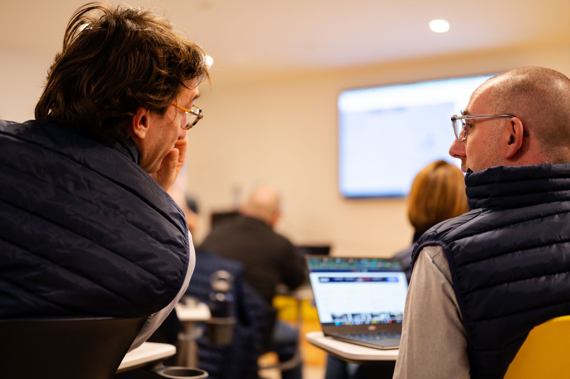 Deux personnes vêtues de doudounes bleu marine sont assises dans une salle de réunion, regardant une présentation, des ordinateurs portables sont à proximité.