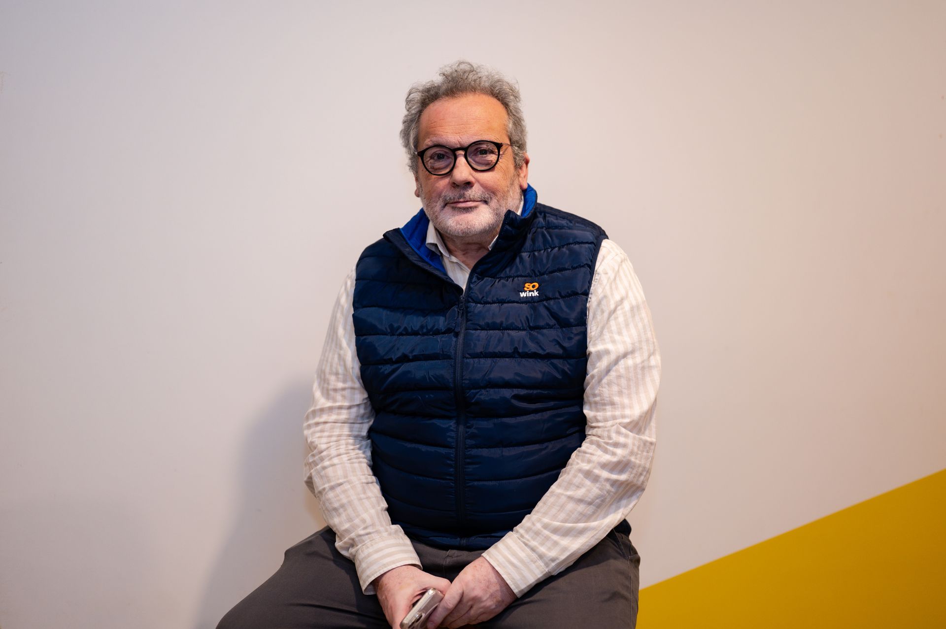 Une personne portant des lunettes et un gilet bleu est assise contre un mur dont un coin est jaune, regardant vers la caméra.